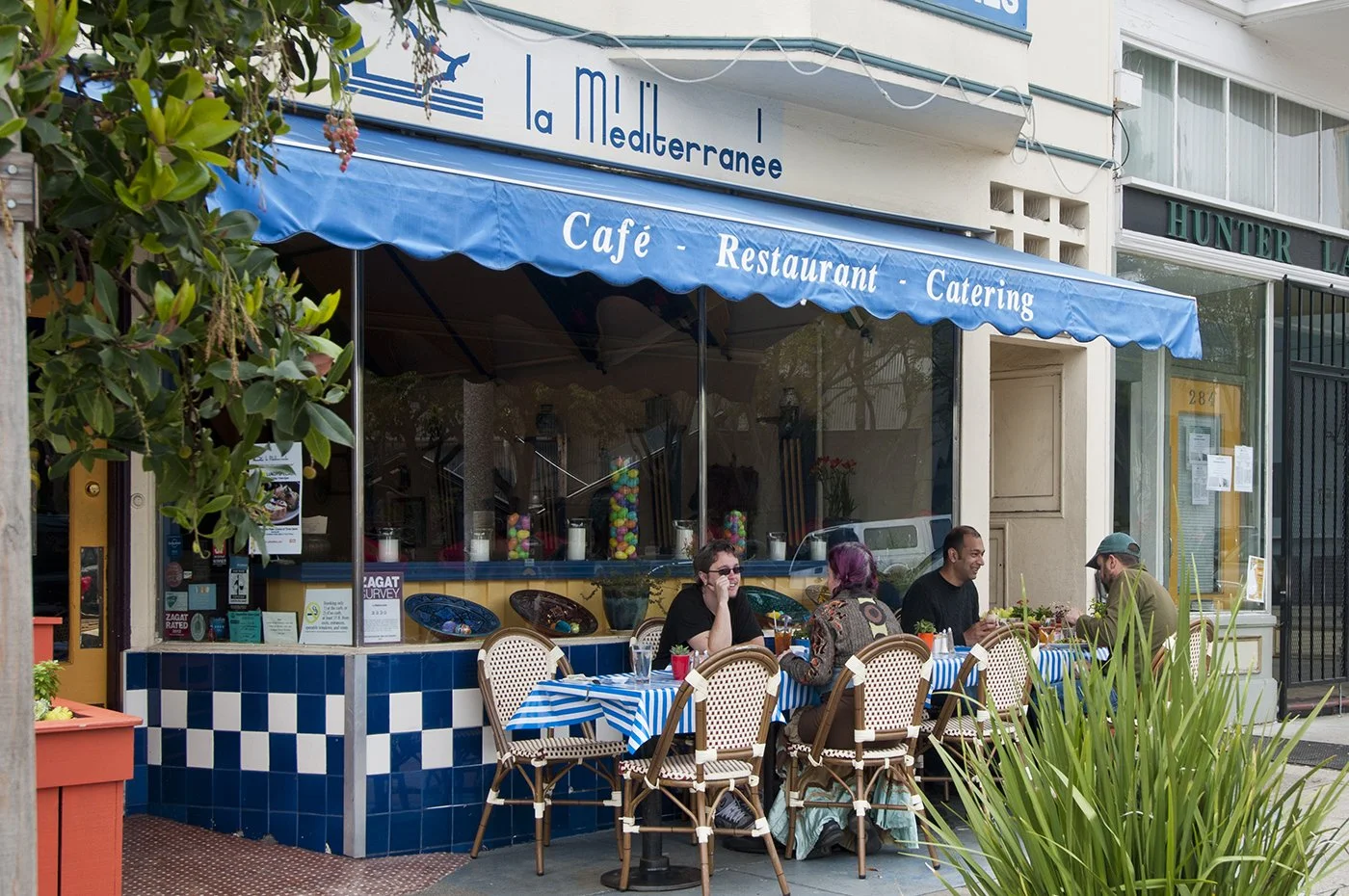 People dining at an outdoor cafe called La Mediterranee with blue and white checkered tablecloths, under a blue awning that reads 'Café - Restaurant - Catering,' in an urban street setting.