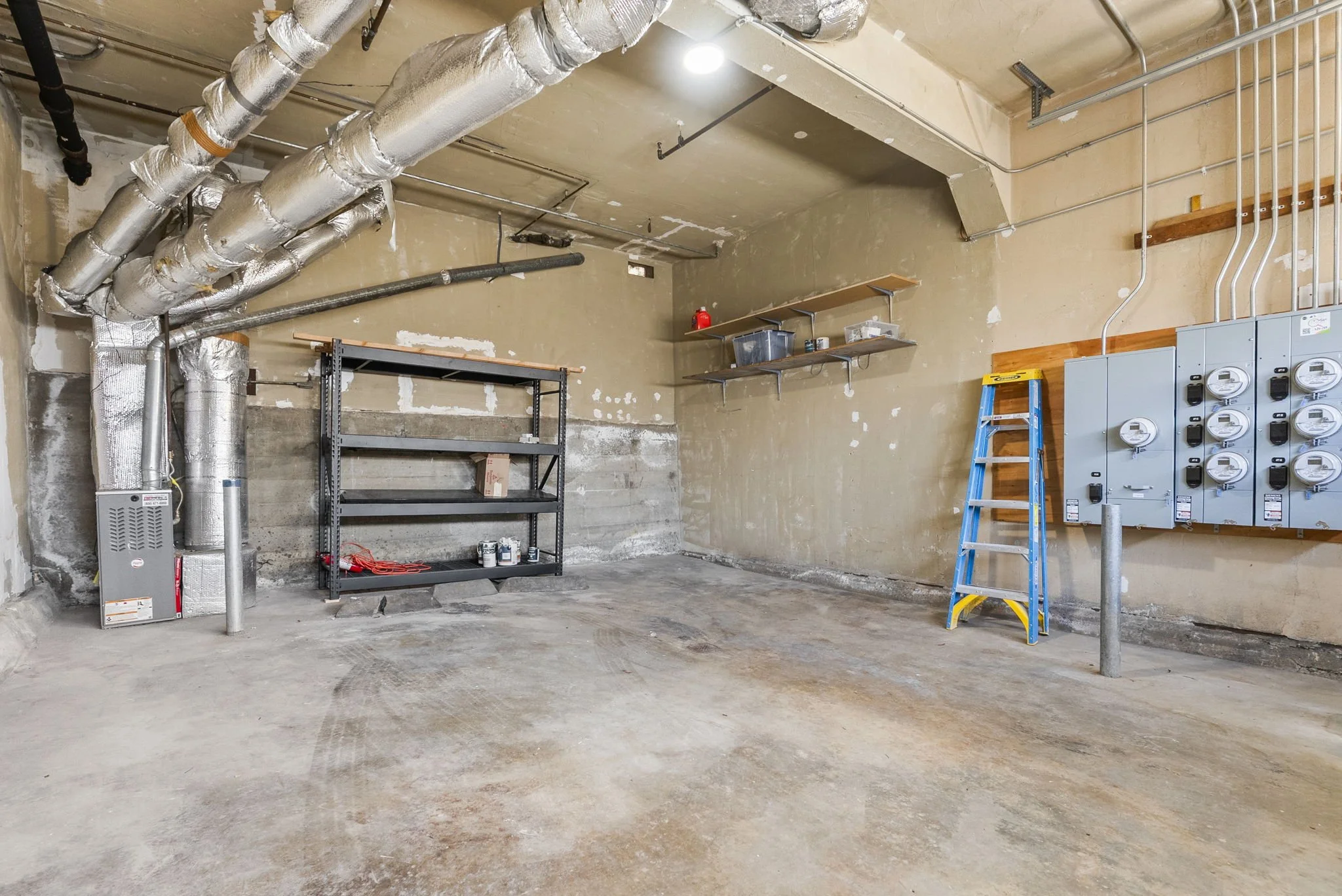 An unfinished garage with exposed ductwork, electrical panels, a ladder, and shelves with various stored items.