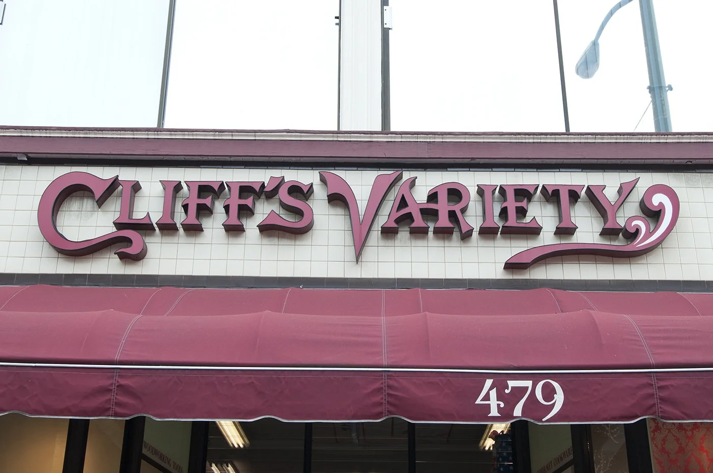 Sign for Cliff's Variety store with large pink lettering and decorative swirls, storefront awning in matching pink, and the building number 479.