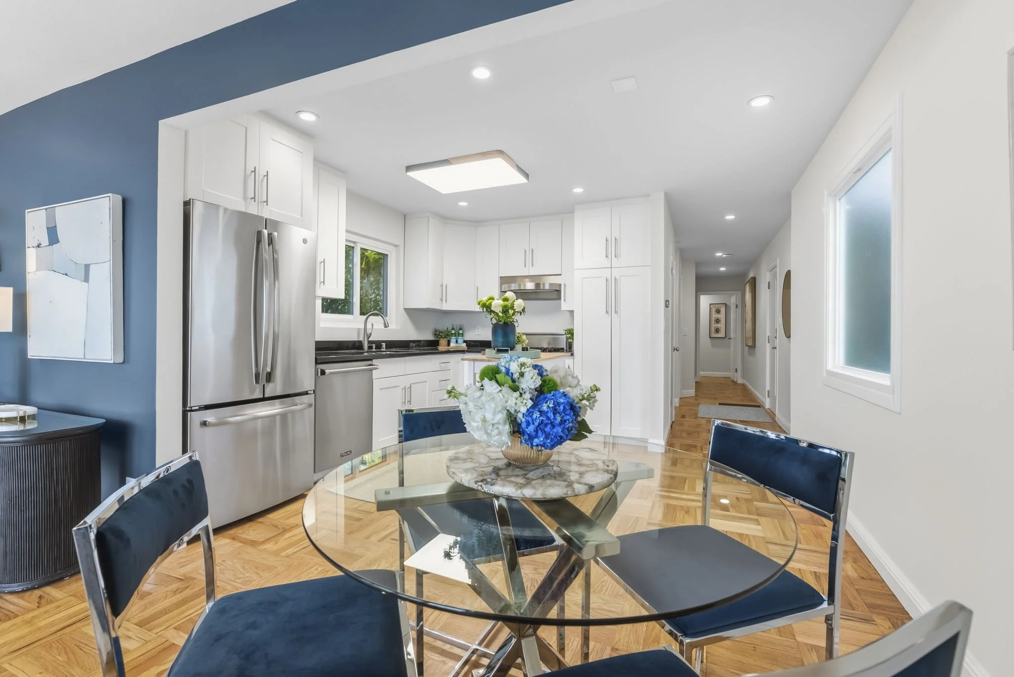A modern kitchen with white cabinets, stainless steel refrigerator, and a round glass dining table with four blue velvet chairs. A bouquet of white and blue flowers is on the table. The kitchen has hardwood floors, a window above the sink, and ceilin