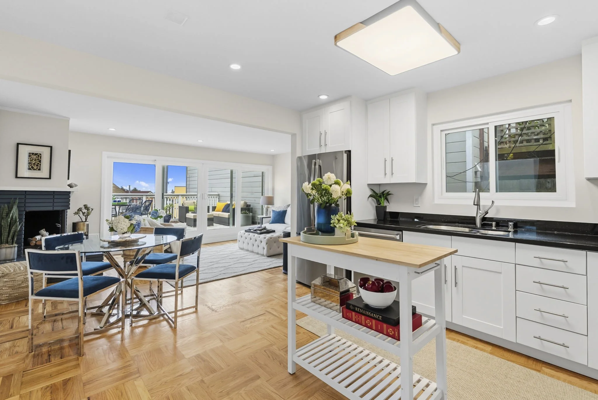 Interior view of a modern, bright kitchen and dining area with white cabinets, stainless steel refrigerator, black countertops, a small island, flowers, and large windows with a patio visible outside.