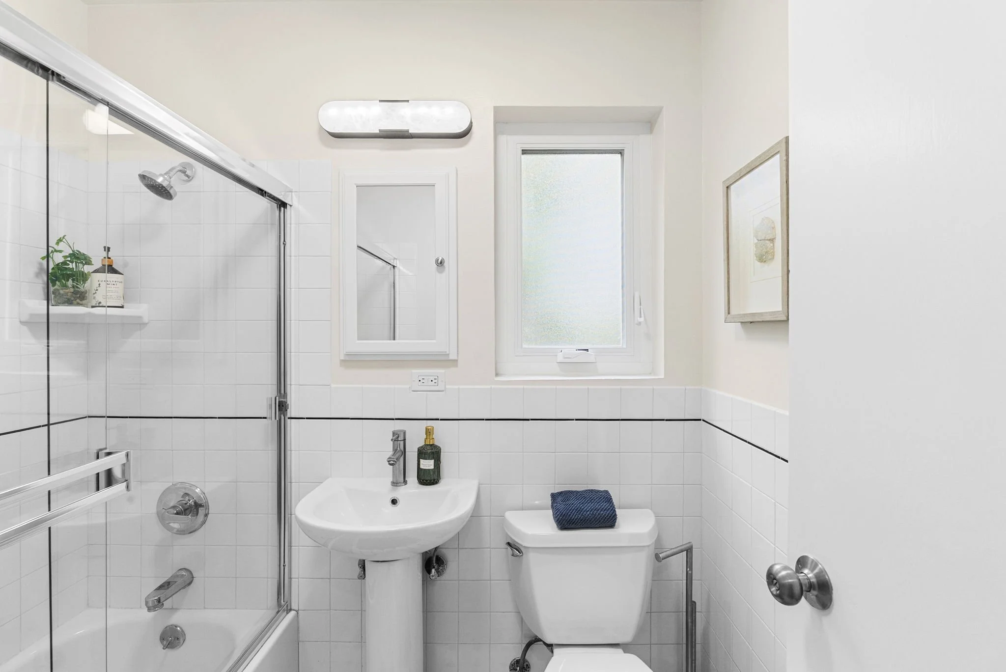 A small bathroom with white tiled walls and a frosted window. It has a shower with glass doors, a white sink with a silver faucet, a green soap dispenser, and a toilet with a dark blue towel on top. There is a wall-mounted mirror and a rectangular li