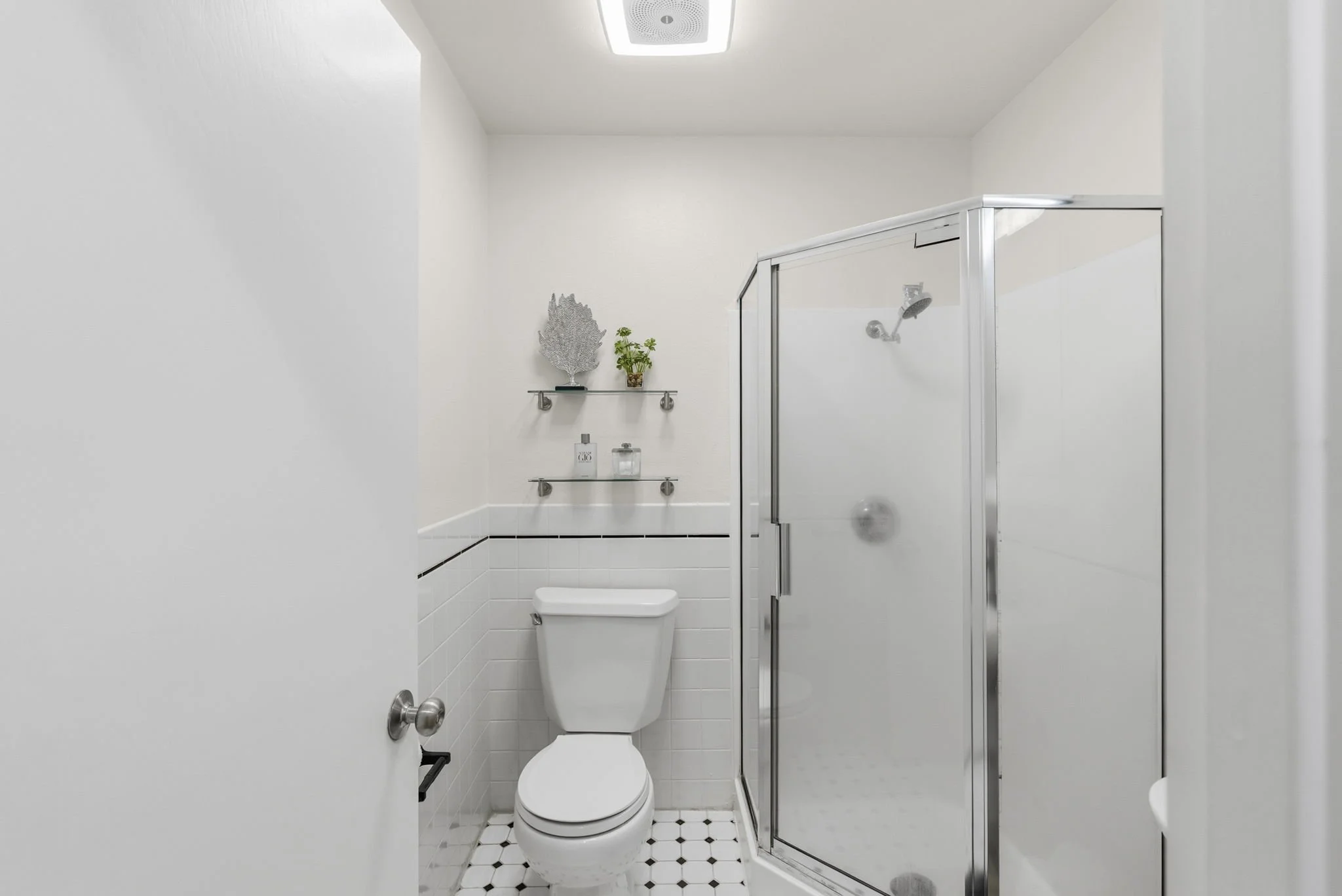A small, modern bathroom with white walls and black and white tile flooring. It contains a white toilet, a glass shower enclosure with a silver frame, and two glass shelves mounted on the wall holding a silver decorative leaf, a small plant, and some