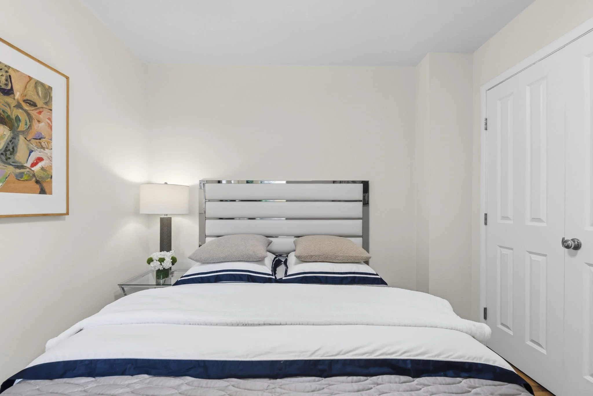 A bedroom with white walls, a bed with blue and white striped bedding, two beige pillows, a nightstand with a vase of white flowers, a table lamp, a framed colorful abstract painting, and a closed white door.