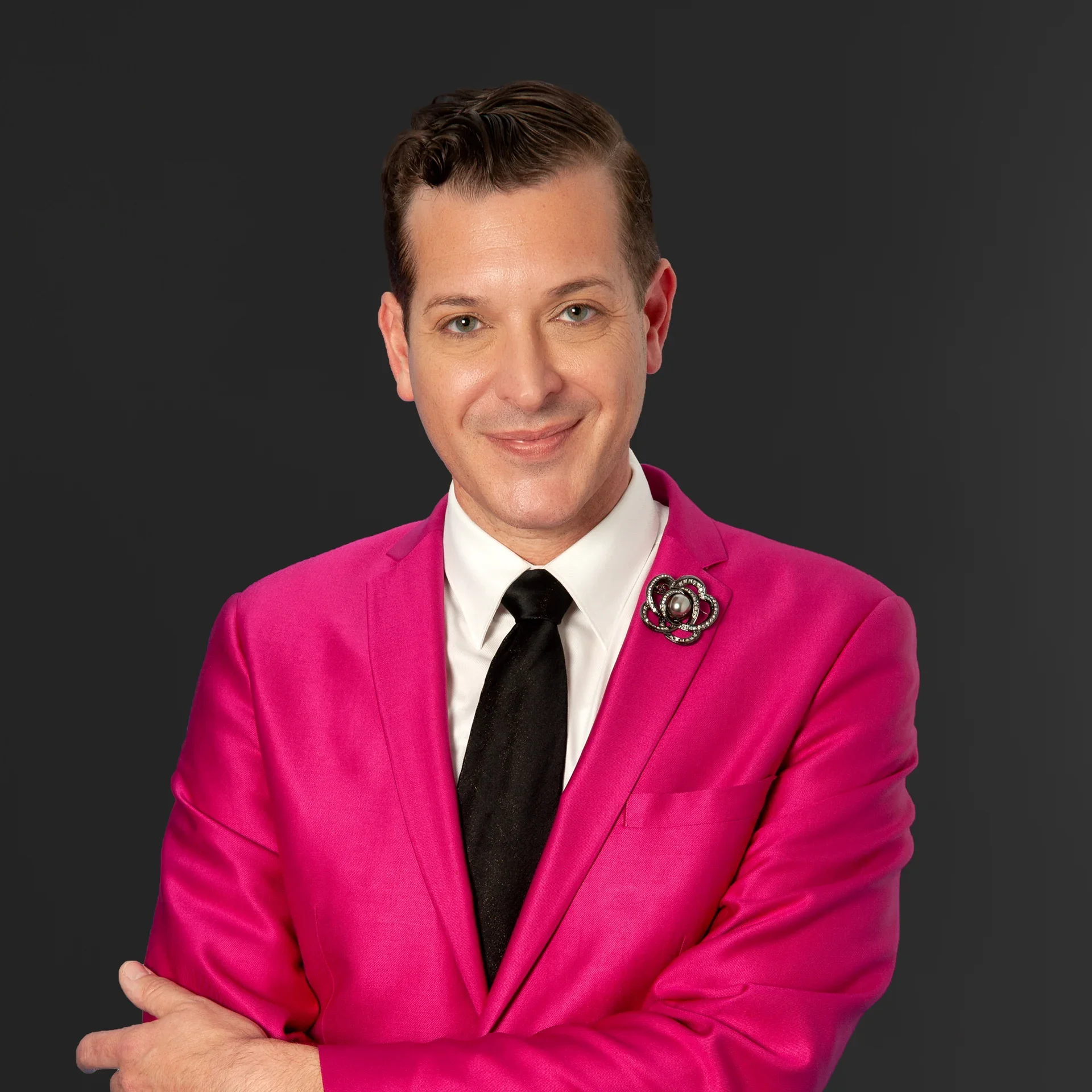 A person wearing a bright pink blazer with a decorative brooch, white shirt, and black tie, smiling slightly, against a dark background.