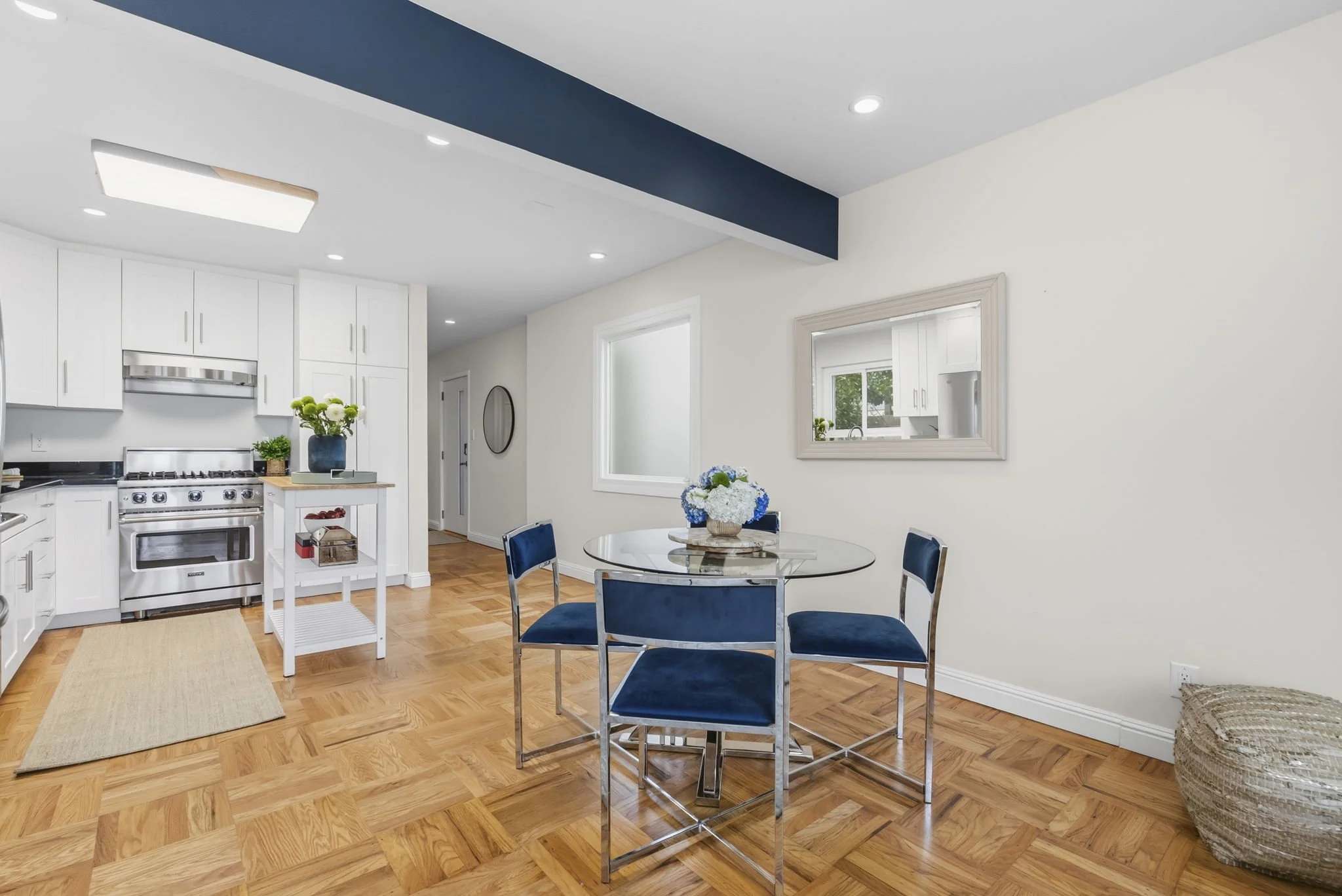 Modern kitchen and dining area with white cabinets, a stainless steel stove, a round glass table with four blue upholstered chairs, and a basket with flowers on the table