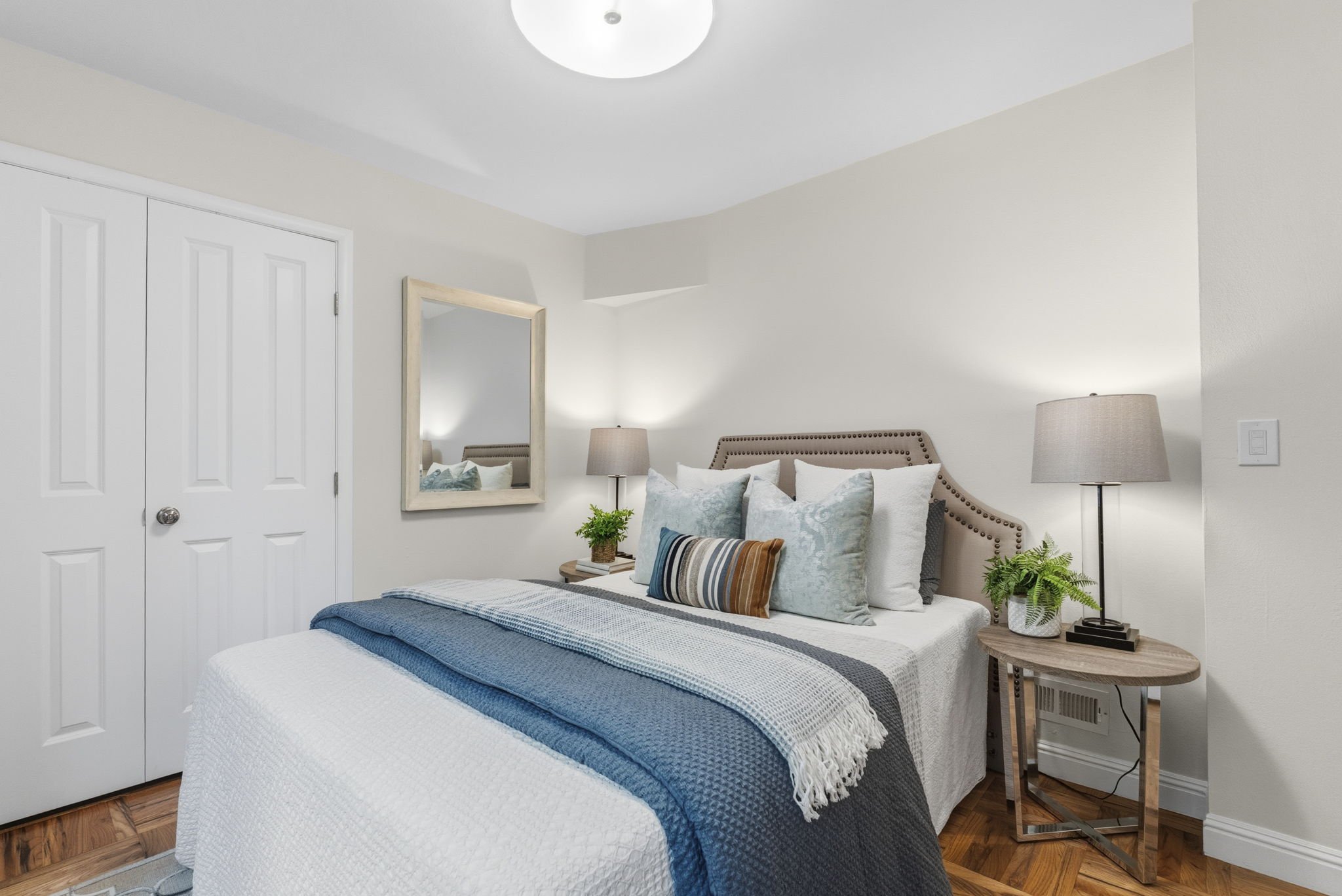 A neatly made bed with white sheets, a blue quilt, and multiple pillows, situated in a bedroom with a beige headboard, two bedside tables with lamps, a large mirror, a potted plant, and a white closet door.