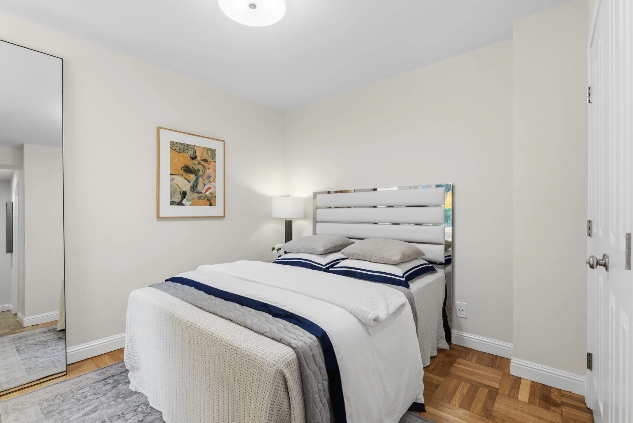 A bedroom with a bed that has white bedding with navy blue accents, two pillows, a nightstand with a lamp, a framed abstract artwork on the wall, a mirrored closet door, and hardwood flooring.