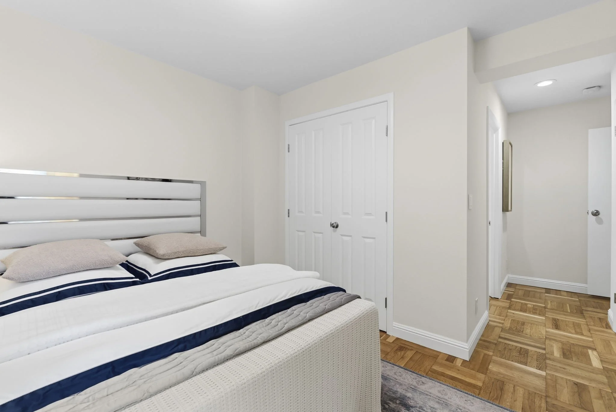 Bedroom with a bed, pillows, white walls, a white door, wooden flooring, and hallway