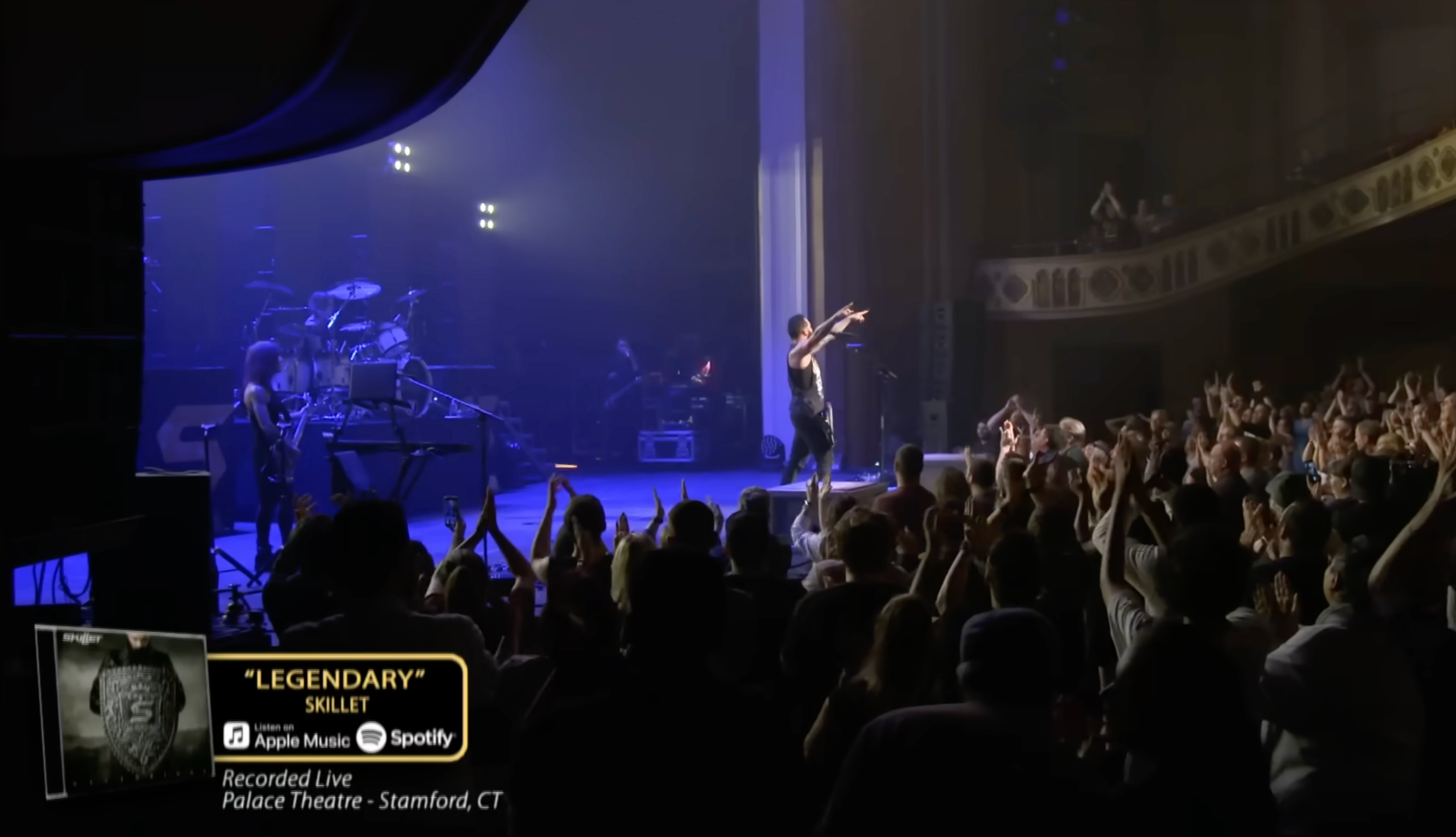 A live concert performance at the Palace Theatre in Stamford, Connecticut. The singer, dressed in black, is on stage with arms outstretched, engaging with the audience. Musicians and instruments are visible on stage. Audience members are reaching towards the performer, cheering. Information about the event, including the song "Legendary" and streaming details, is displayed on a screen.