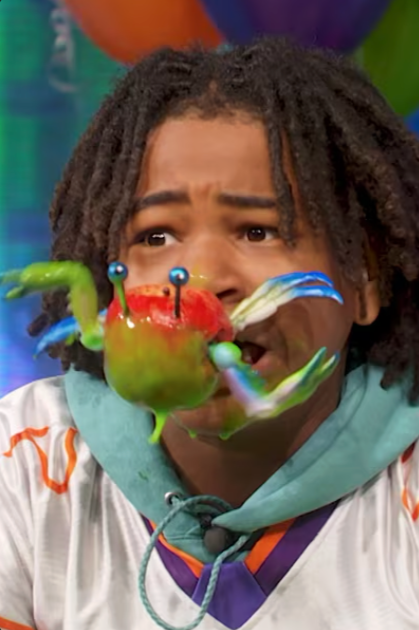 A person with dreadlocks and a teal shirt is looking worried while wearing a colorful, toy crab on their face.