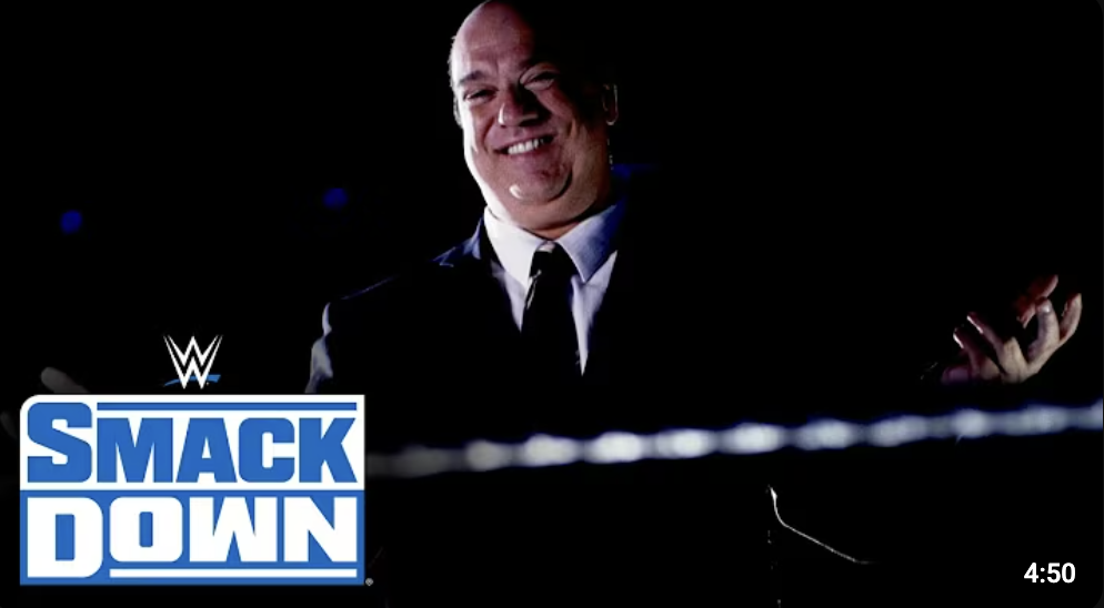 A man in a suit smiling in a dark setting with the WWE SmackDown logo in the bottom left corner.