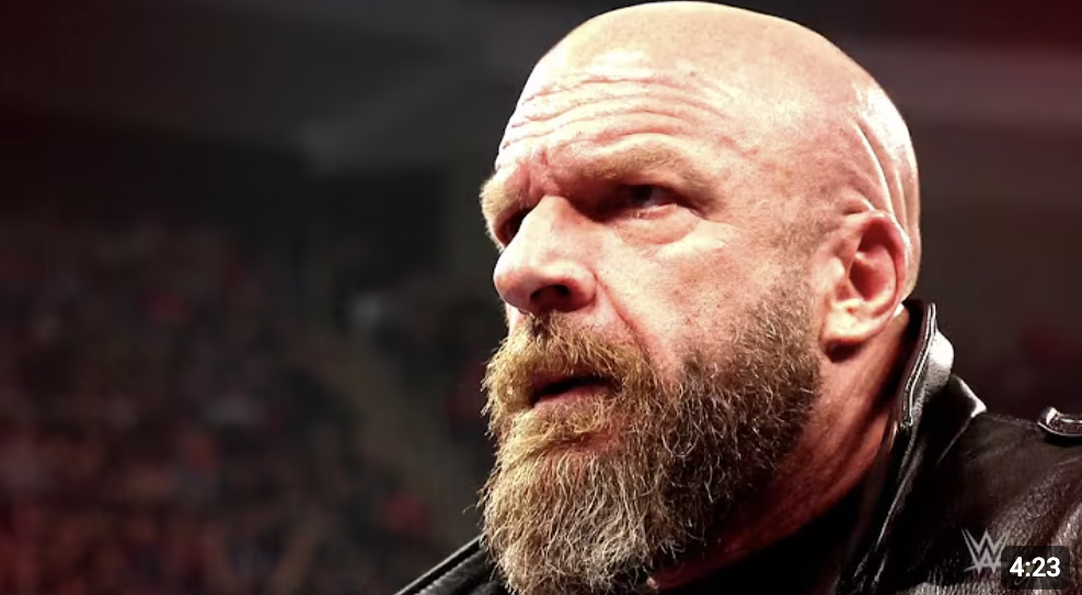 Close-up of a bald man with a full beard, wearing a black leather jacket, in a wrestling arena.