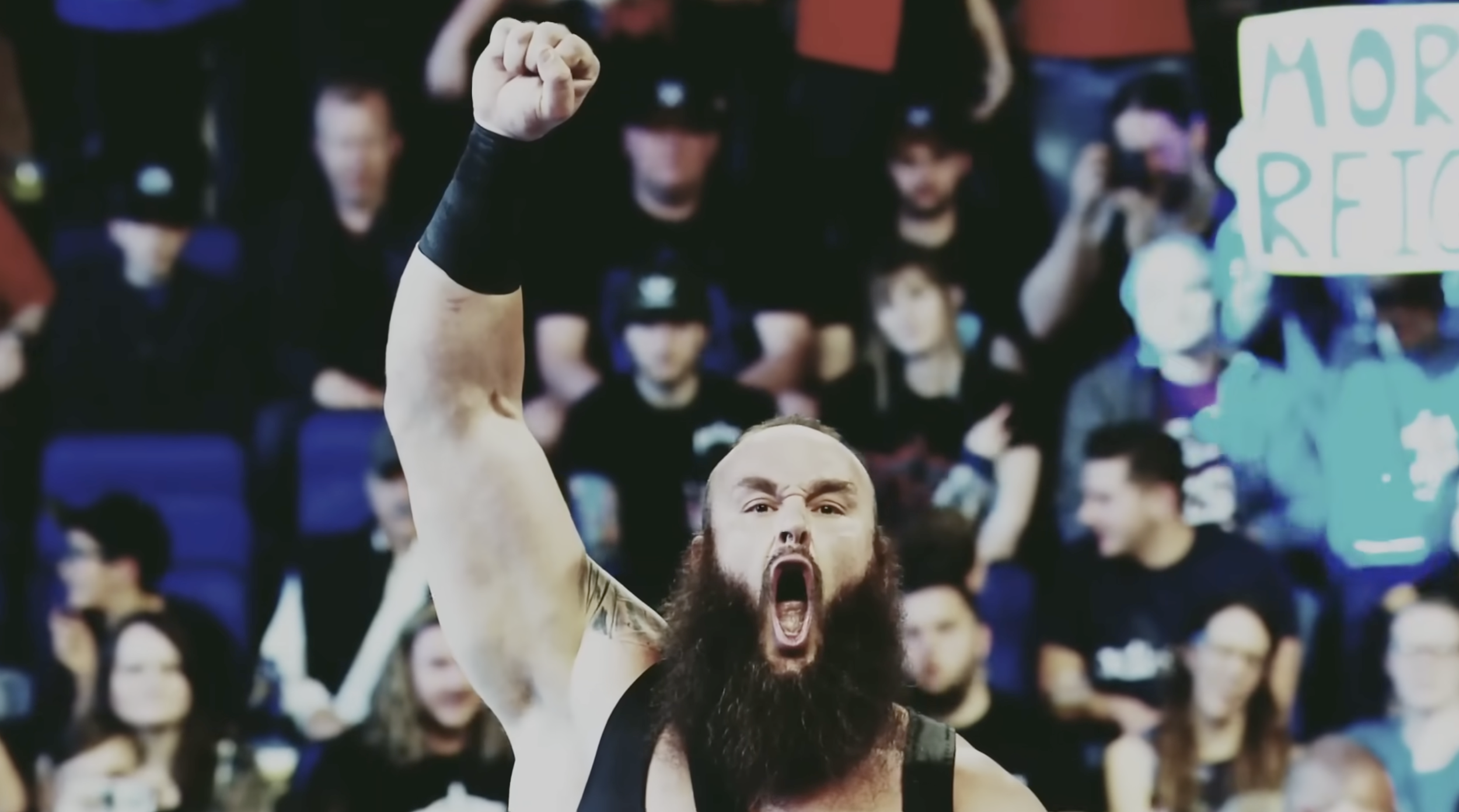A man with a beard and tattoos on his arm, with his mouth open, raising his fist in victory during a sports event, with a crowd in the background holding up signs and phones.