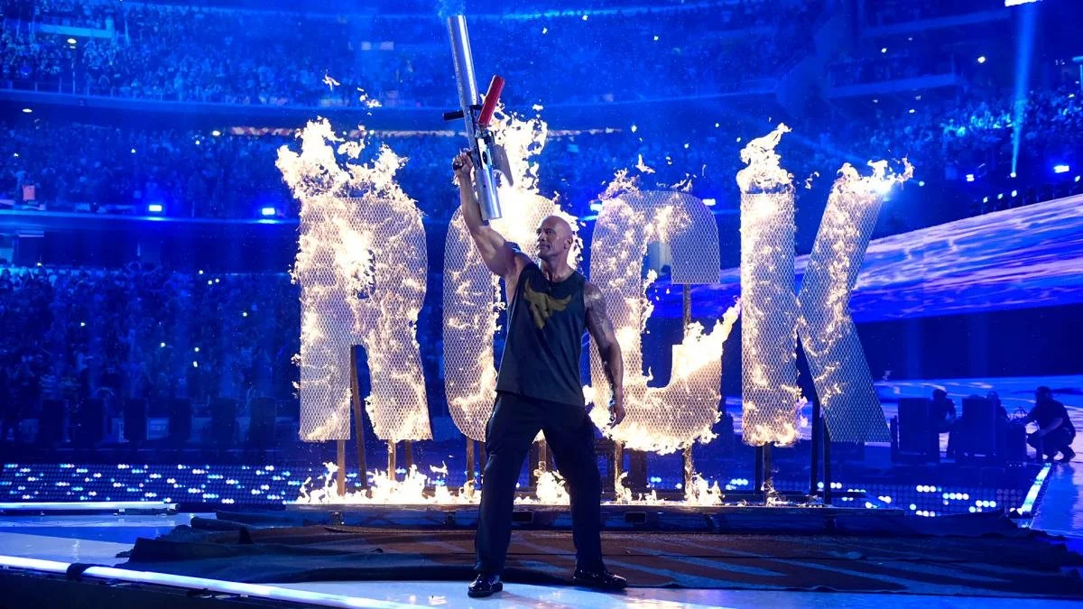 A man holding a gun above his head on a stage with large burning letters behind him.