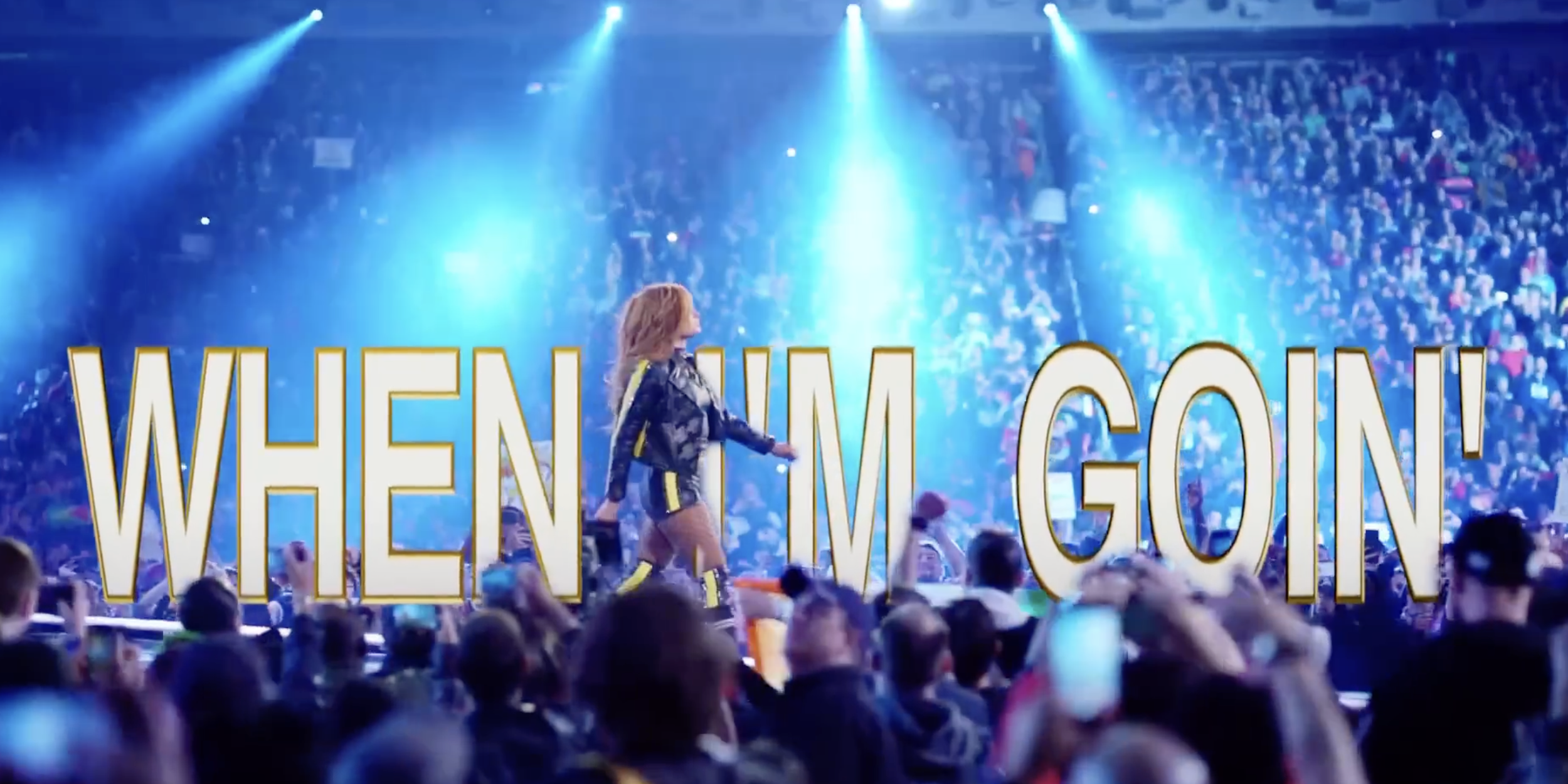 A performer on stage in front of a large crowd, with bright blue lighting and the words "WHEN I GO" displayed in large yellow letters across the scene.