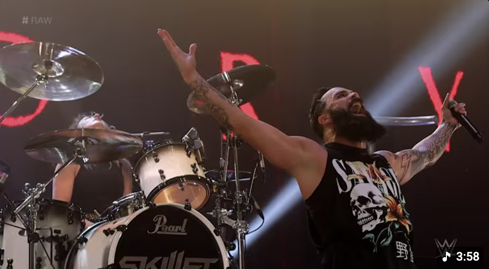 A male drummer with long hair, beard, and tattoos on his arms, playing a white Pearl drum set on stage. A female drummer with blonde hair is partially visible behind him. The man is holding a microphone in his right hand and has his arms raised, celebrating on stage with a large red LED screen in the background.