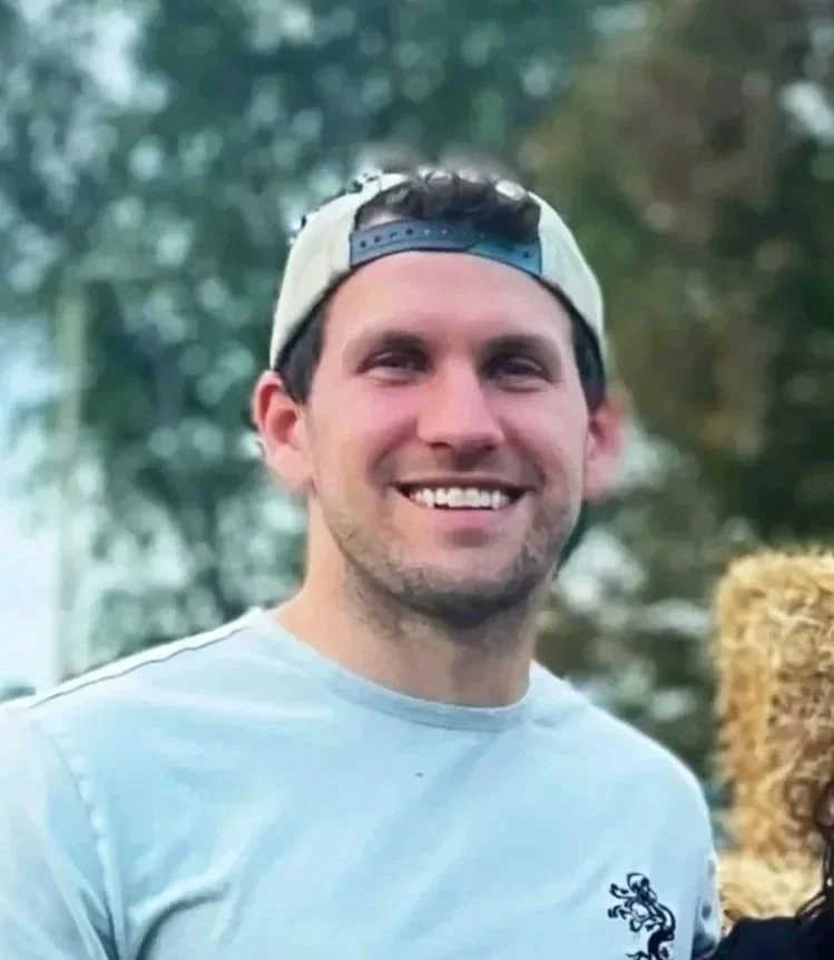 Smiling young man wearing a light gray t-shirt and a white and dark blue baseball cap backward outdoors with trees in the background.