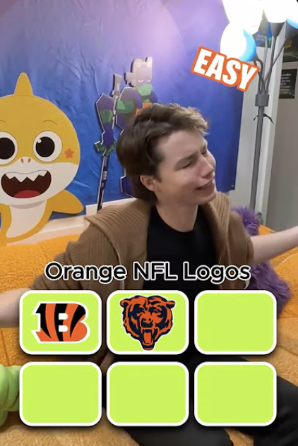 Young person with short brown hair and brown sweater, crying or distressed, sitting on an orange and purple plush surface, in front of a colorful backdrop with cartoon characters and NFL logos on cards.