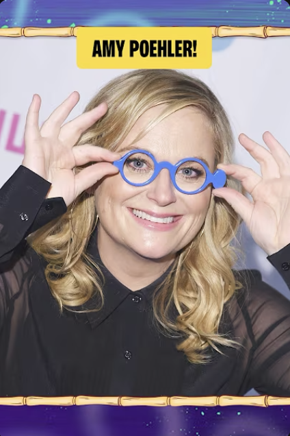 Woman with wavy blonde hair and a black blouse wearing novelty blue glasses, smiling and adjusting the glasses with both hands, against a backdrop with the text 'Amy Poehler!'