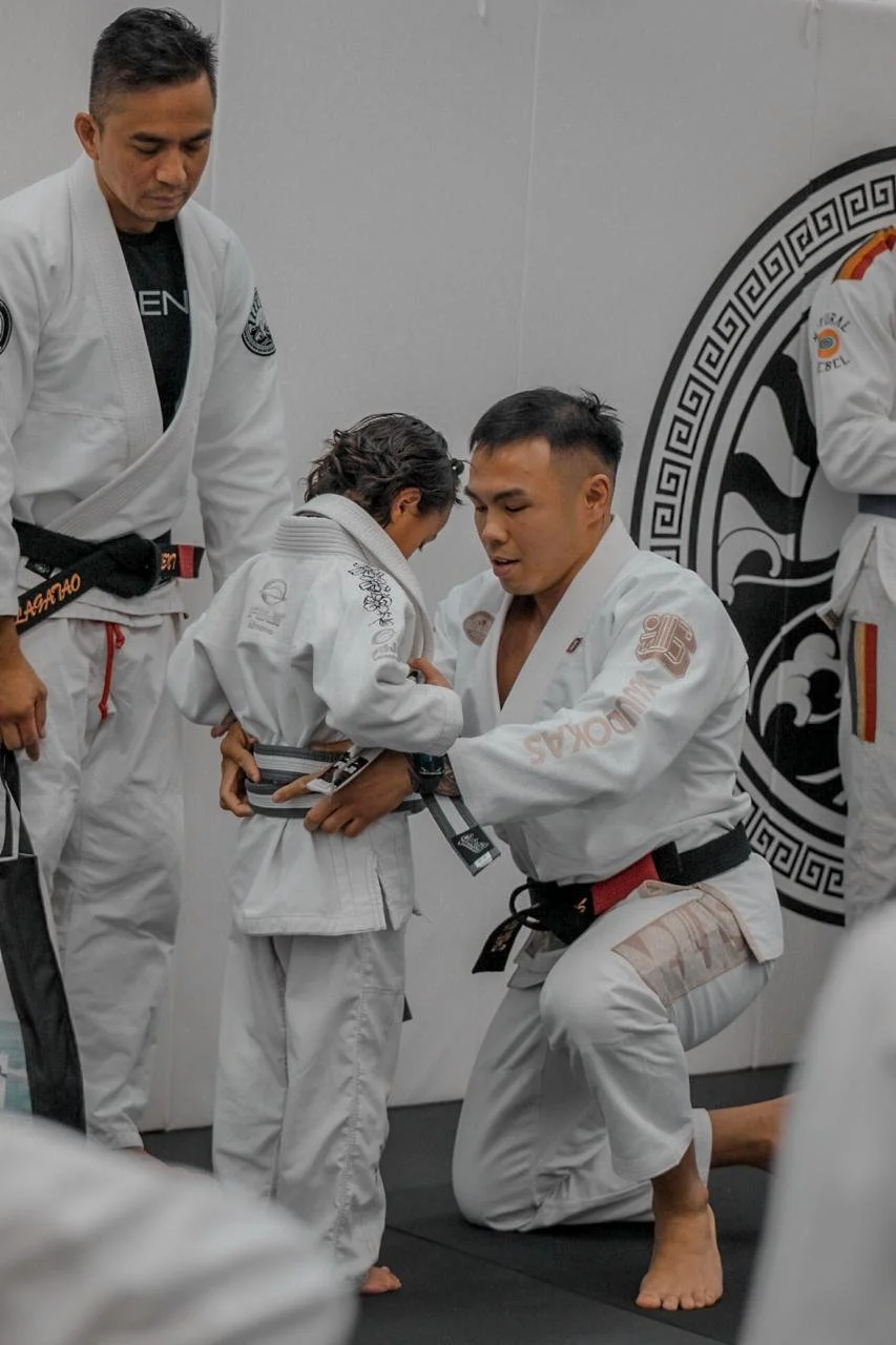 A Brazilian Jiu-Jitsu instructor kneeling and assisting a young girl with her gi in a martial arts class, surrounded by other students in gis.