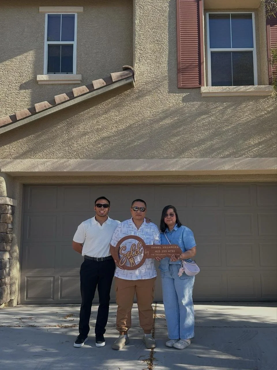 Three people standing in front of a garage door, holding a large orange sign that says "Sold" with a name and phone number. Two men and one woman, all wearing sunglasses and casual outfits.