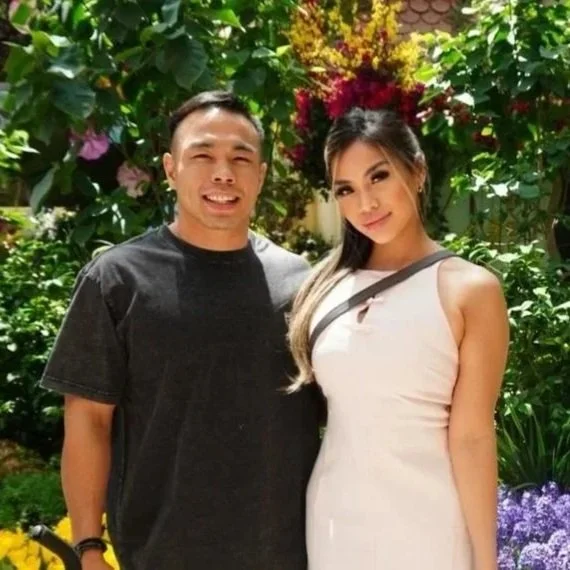 A man and woman smiling outdoors surrounded by lush green plants and colorful flowers.