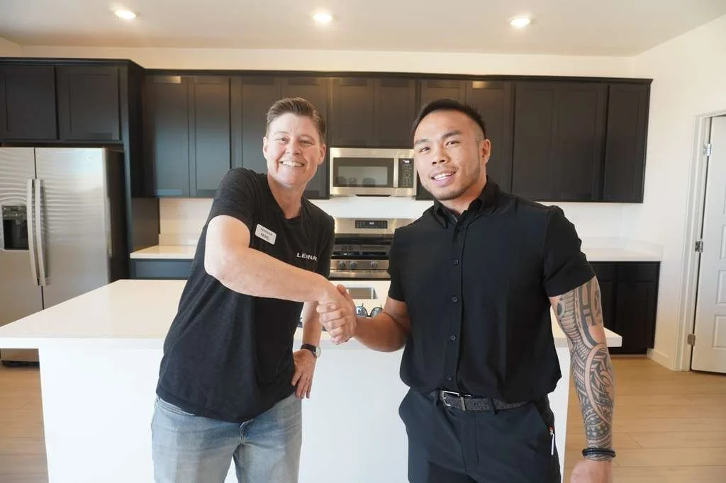 A woman and a man shake hands in a modern kitchen. The woman is smiling and wearing a black shirt, and the man has a tattooed arm and is wearing a black shirt.