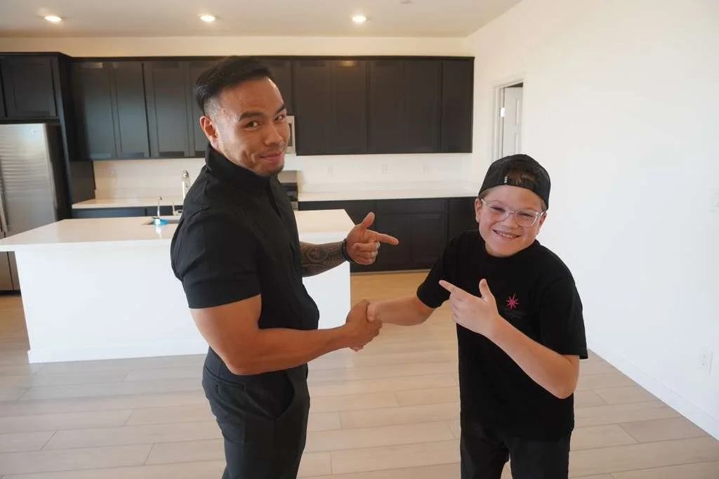 Two men shaking hands and pointing at each other in a modern kitchen with black cabinets and white walls.
