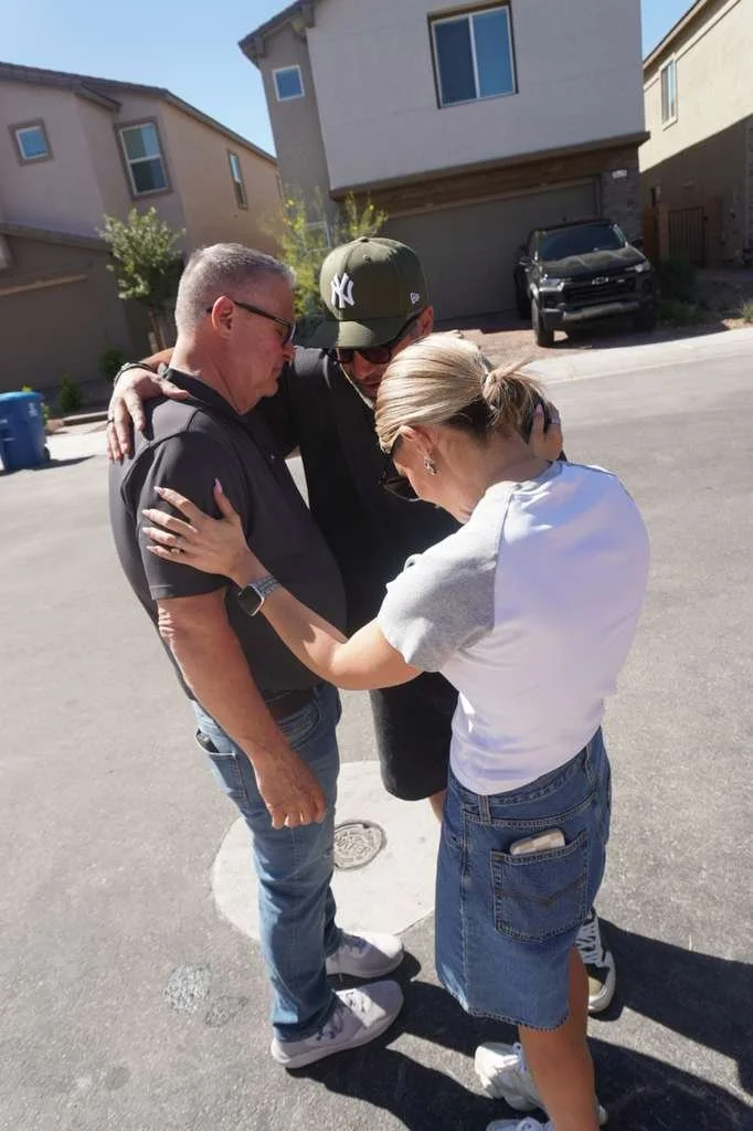 Three people, two men and one woman, are embracing in a tight hug on a residential street. The woman is wearing a white t-shirt and denim shorts, while the men are wearing dark clothing and baseball caps. The scene is outdoors with houses and a car p