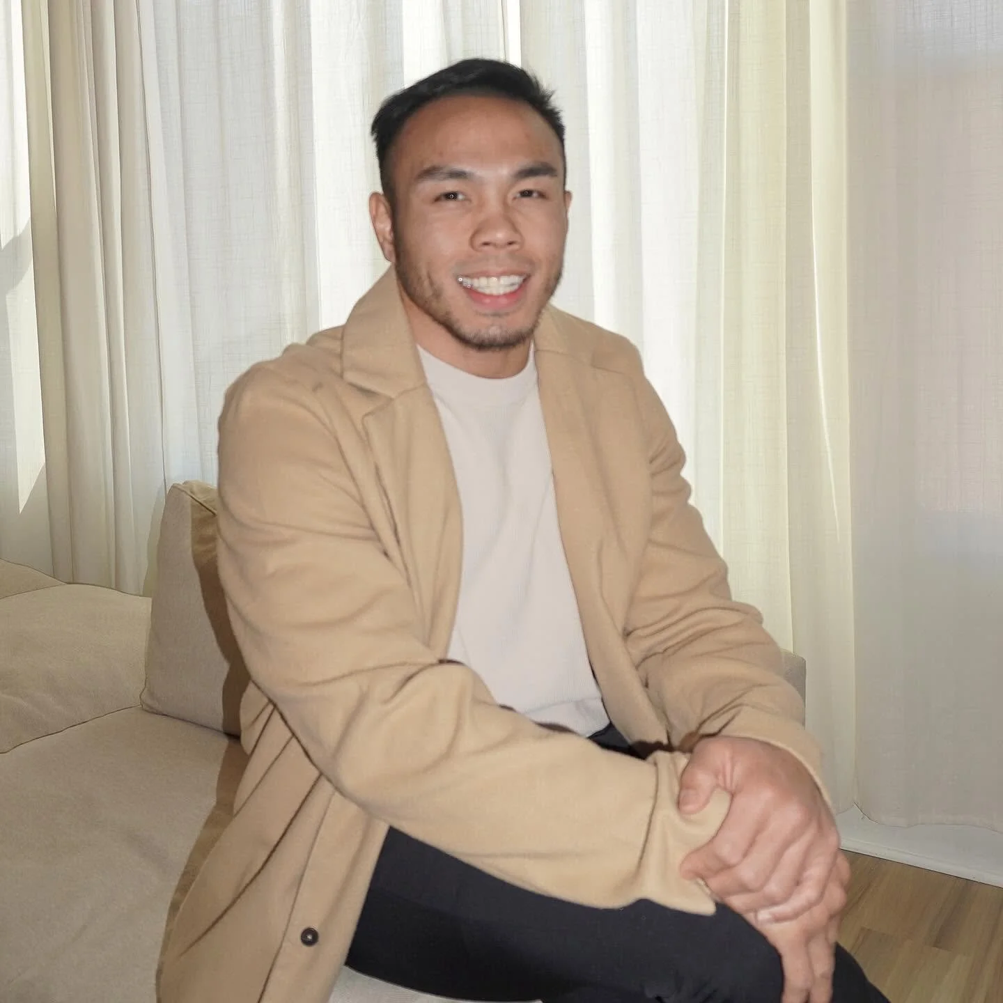 A smiling man sitting on a beige sofa with a beige jacket and white shirt, in front of white curtains.