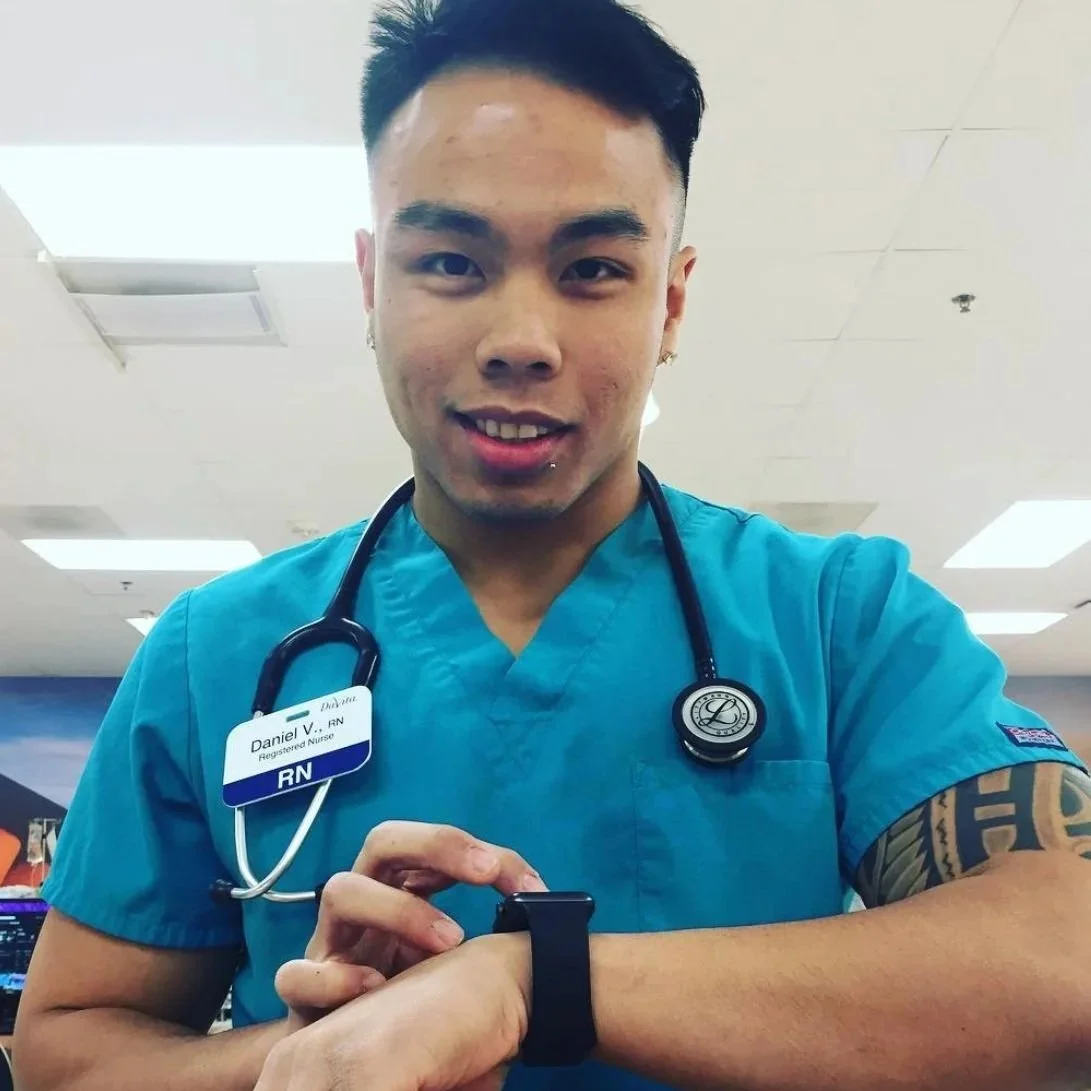 A smiling young male nurse or healthcare worker in blue scrubs, wearing a stethoscope around his neck, looking at the camera indoors.