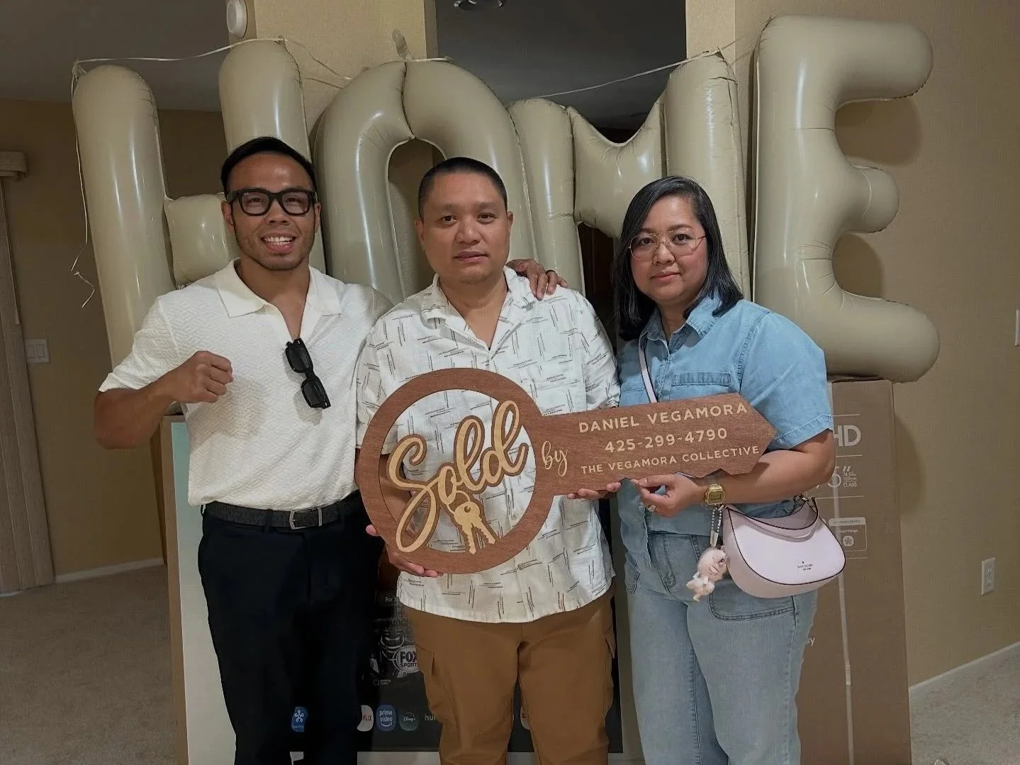 Three people standing together indoors, holding a wooden sign that says 'Sold by Daniel Vegamora 425-299-4790 THE VEGAMORA COLLECTIVE,' in front of large beige balloons spelling 'HOME' and a beige wall.