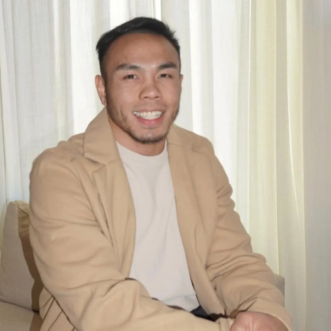 A smiling man with short dark hair and facial hair, wearing a beige jacket and white shirt, sitting in front of light-colored curtains.