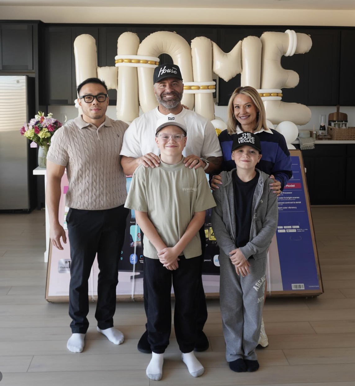 Group of five people standing in front of large balloon letters spelling 'HOME' at a celebration or event.