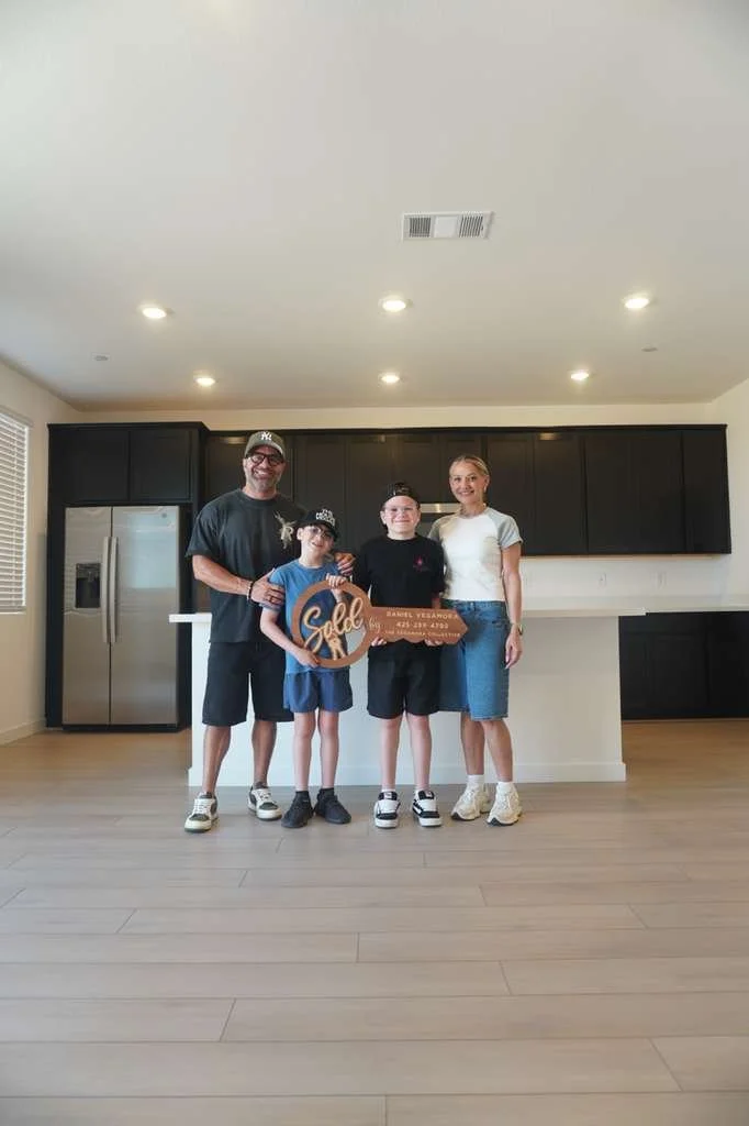 A family of four standing inside a new house with an empty kitchen. They are smiling, holding a sign that says "Sold," indicating they are celebrating buying a new home.