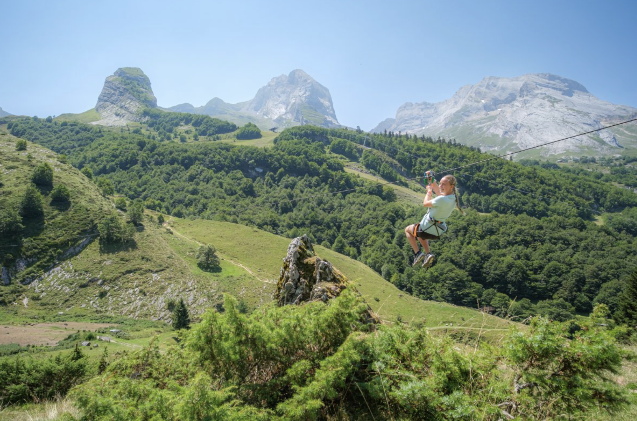 Une jeune fille fait de la descente en tyrolienne dans un paysage de montagnes verdoyantes avec des rochers imposants et un ciel clair.