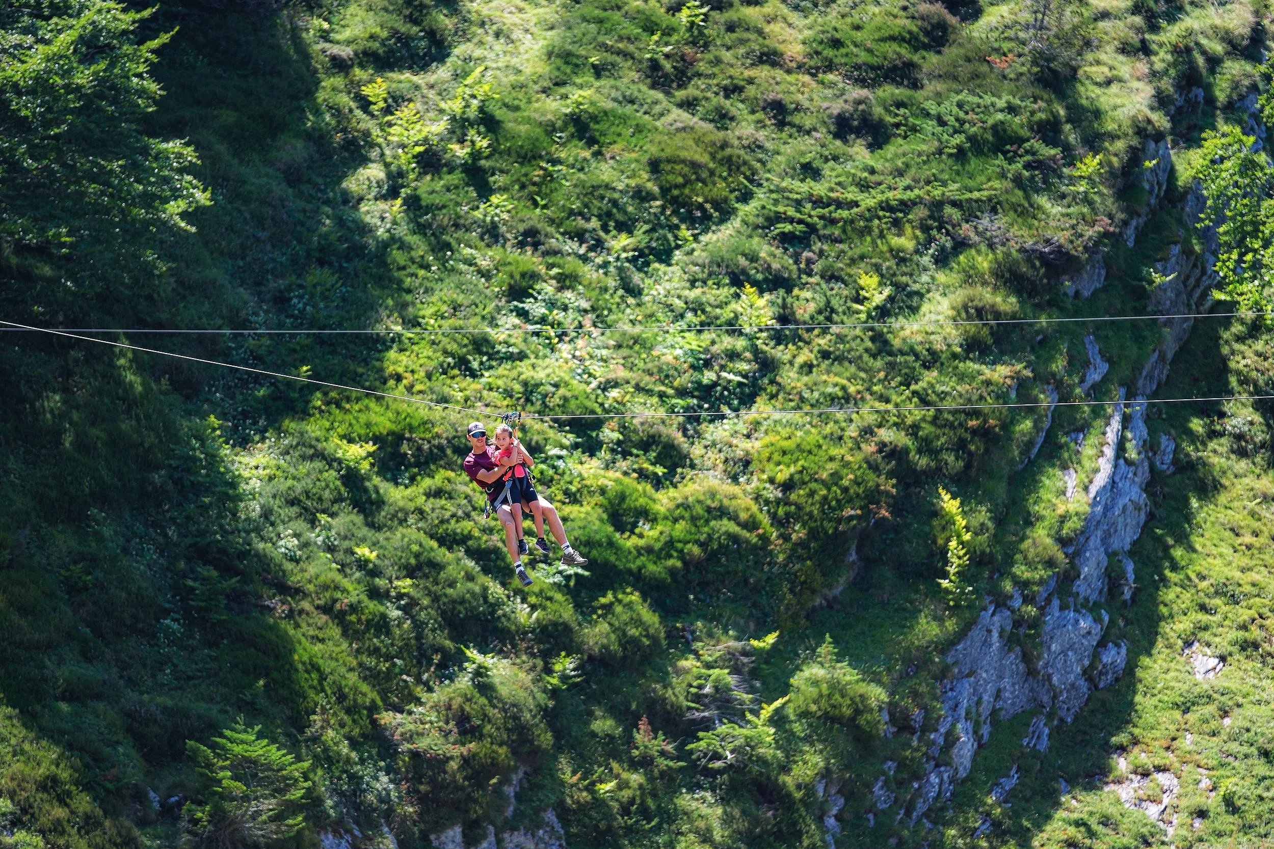 Deux personnes, un adulte et un enfant, font une descente en Tyrolienne au-dessus d'une forêt verte dense en montagne.