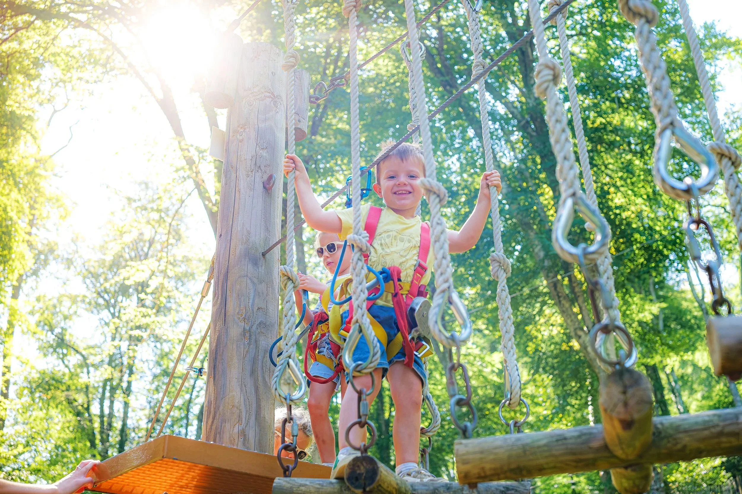 Deux enfants jouent sur une structure de jeu en plein air entourée d'arbres et de lumière solaire. L'un d'eux, un garçon, sourit tout en montant sur une plateforme suspendue avec des cordes.