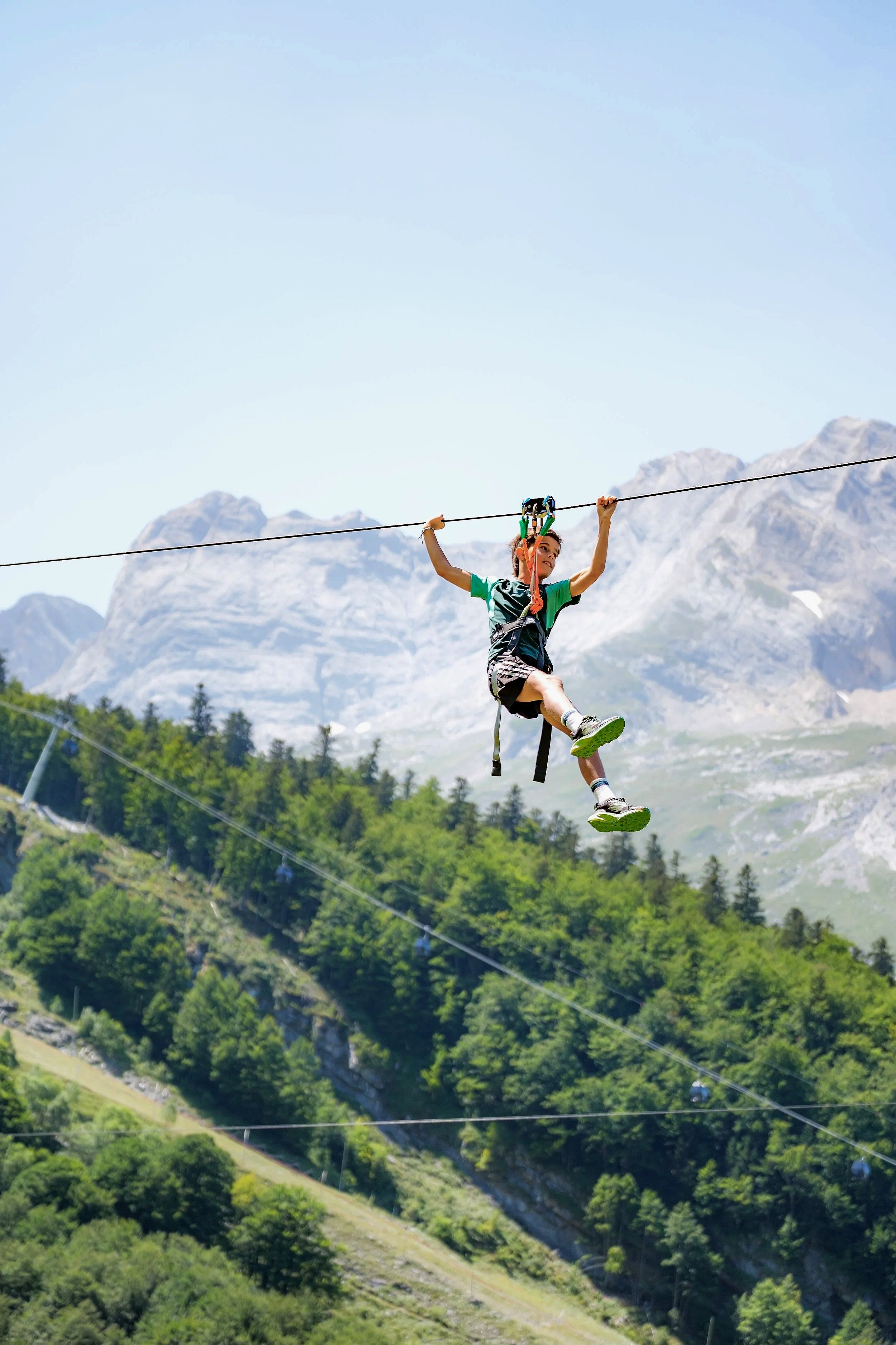 Une personne en tenue de sport circule sur une tyrolienne au-dessus d'une vallée verdoyante avec des montagnes en arrière-plan, sous un ciel clair.