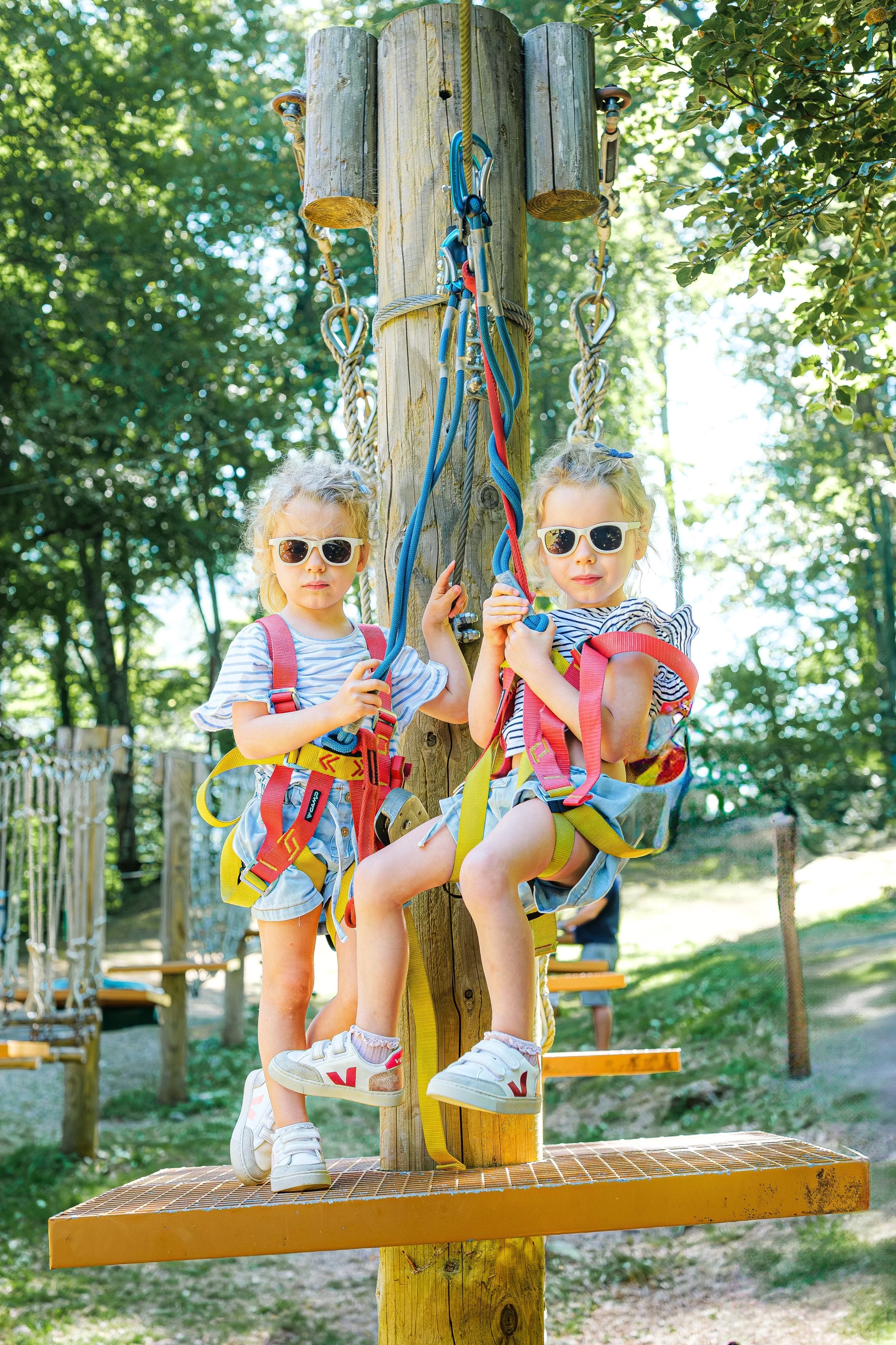 Deux jeunes filles portant des lunettes de soleil et un harnais d'escalade se tiennent sur une plateforme en métal d'un parcours d'accrobranche dans un parc arboré, avec des arbres en arrière-plan.