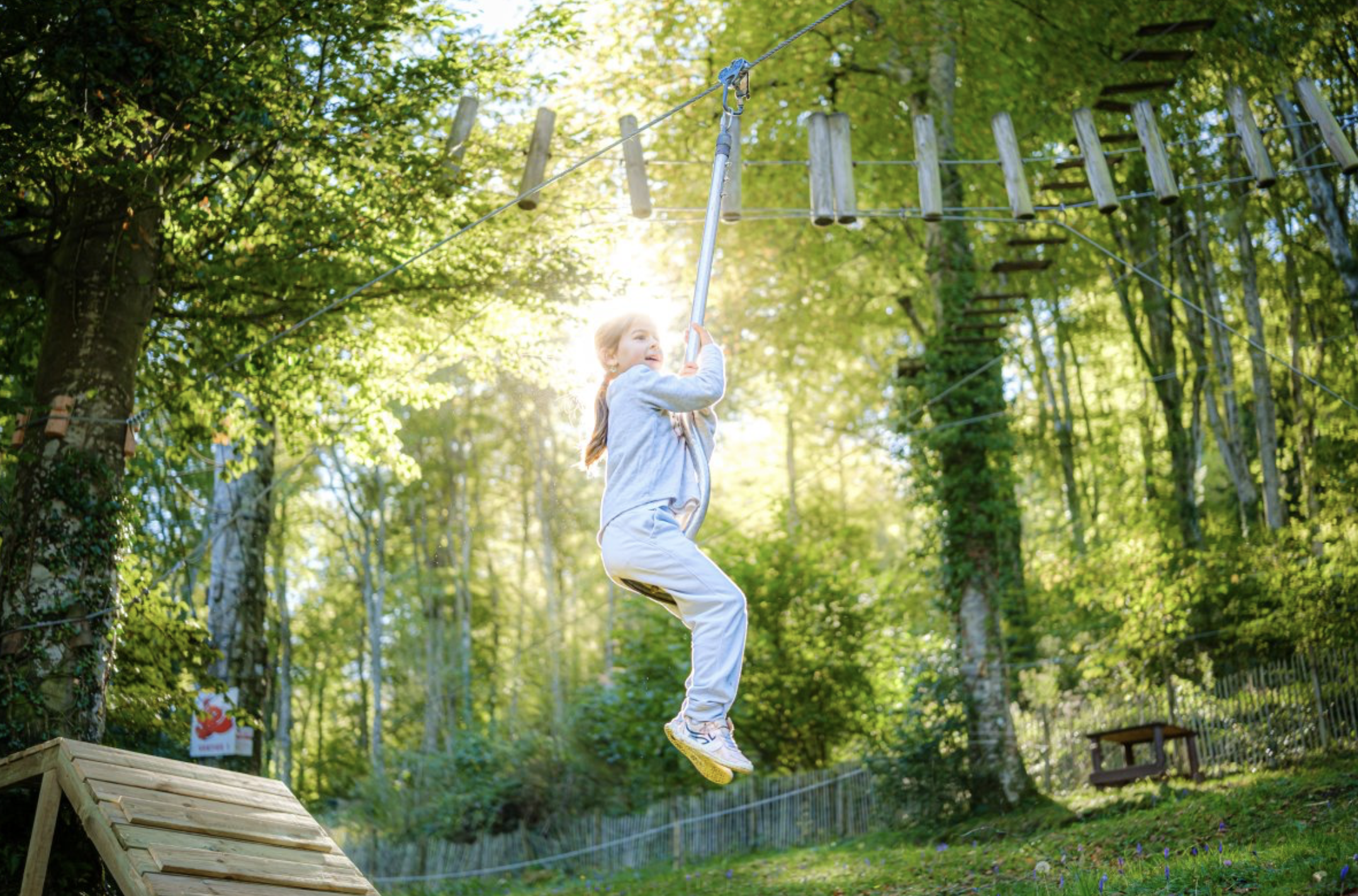 Une jeune fille en vêtements décontractés, suspendue à une ligne de zipline en forêt lors d'une activité de plein air, avec un fond d'arbres et de soleil filtrant à travers les feuilles.