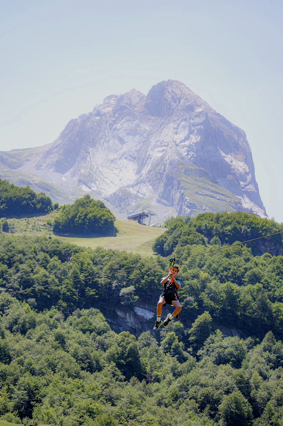 Un garçon faisant de la tyrolienne dans un paysage montagneux verdoyant avec une grande roche en arrière-plan.