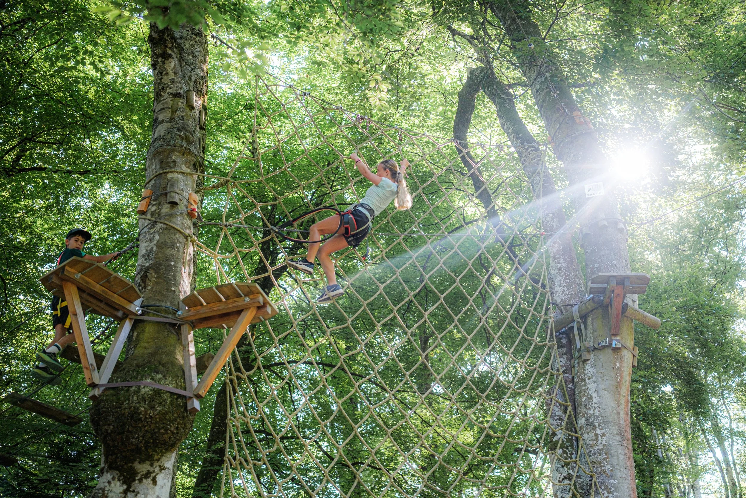 Enfants jouant sur un parcours d'accrobranche dans une forêt avec des arbres verts. U ne fillette traverse un filet suspendu en hauteur, une autre enfant se tient sur une plateforme en bois, tous portent un harnais de sécurité.