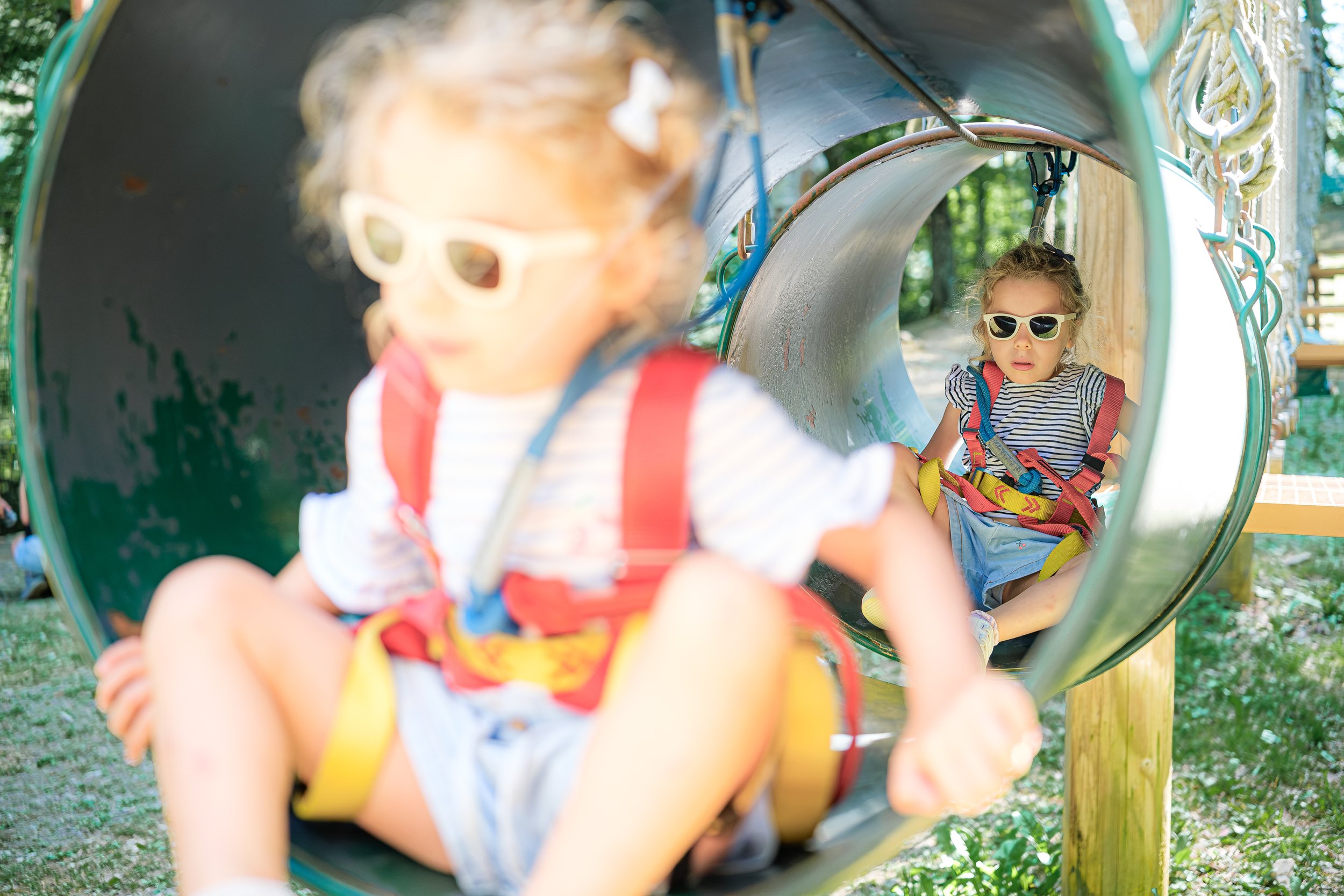Deux enfants jouent dans un tunnel de jeu en extérieur, portant des gilets de sécurité et des lunettes de soleil, parmi des arbres.