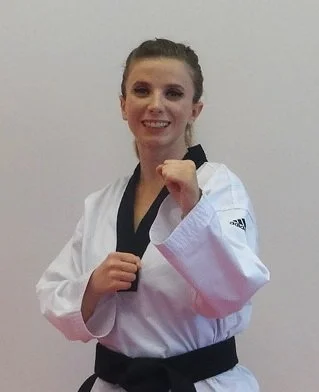 A smiling woman in a taekwondo uniform posing with fists up in a kicking stance.