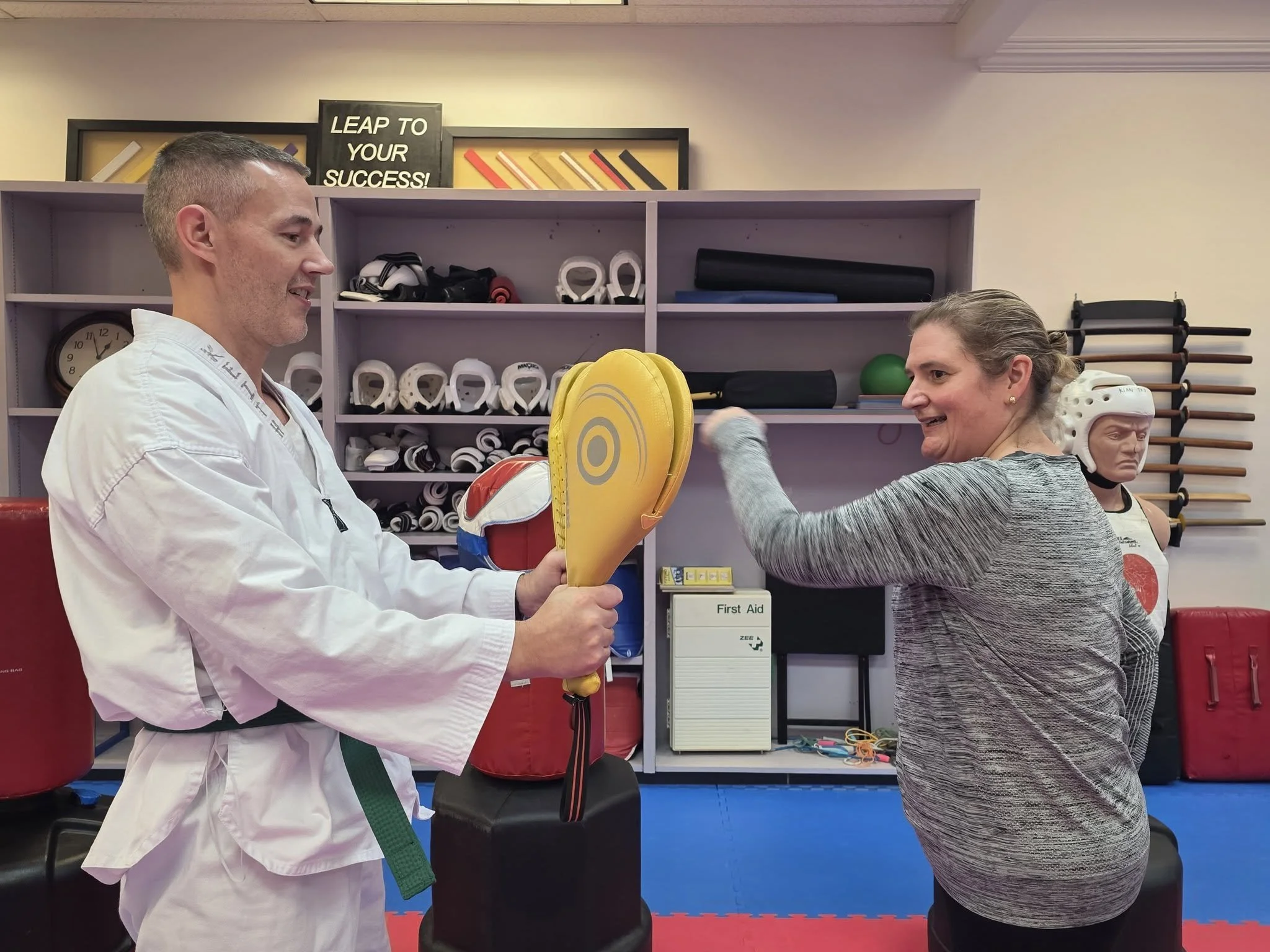 A student in a white uniform holding targets for a a woman in a gray sweater so that she can punch them.