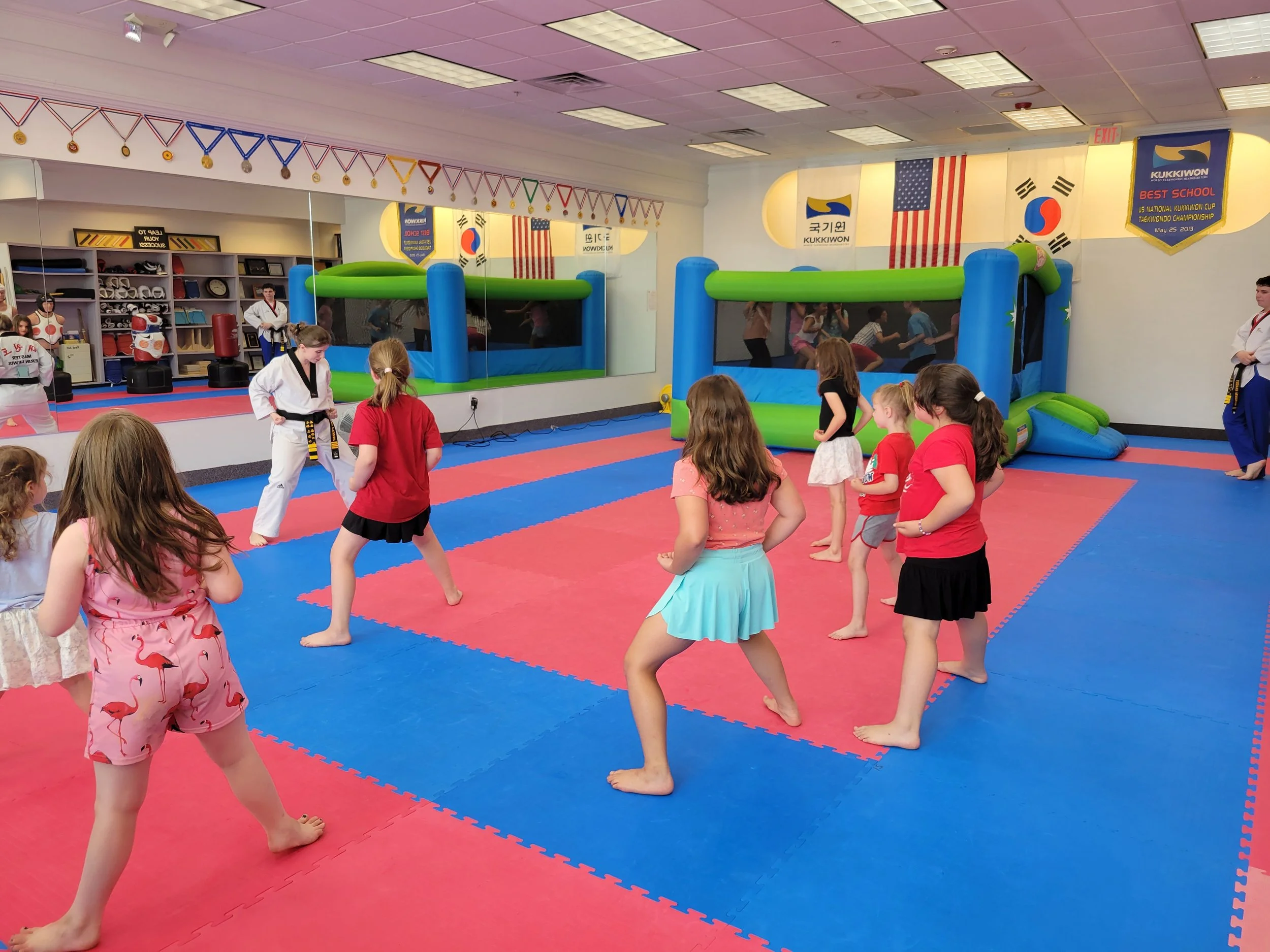 Children in a martial arts class practicing Taekwondo with a Master in a dojang that has flags and banners, including the South Korean flag and U.S. flags.
