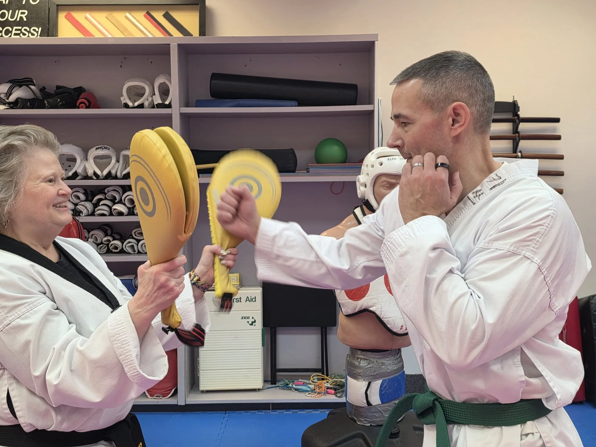 Two martial artists in white doboks practicing in a dojang. The woman is holding foam training pads, and the man is striking at the pads with a fist.