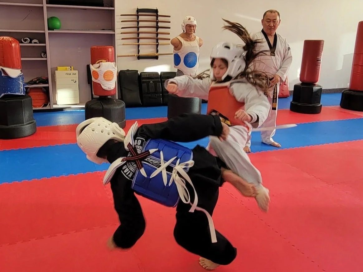 Two students practicing taekwondo with protective headgear in a dojang, one student executing a kick while the other falls down with a master referring in the background.